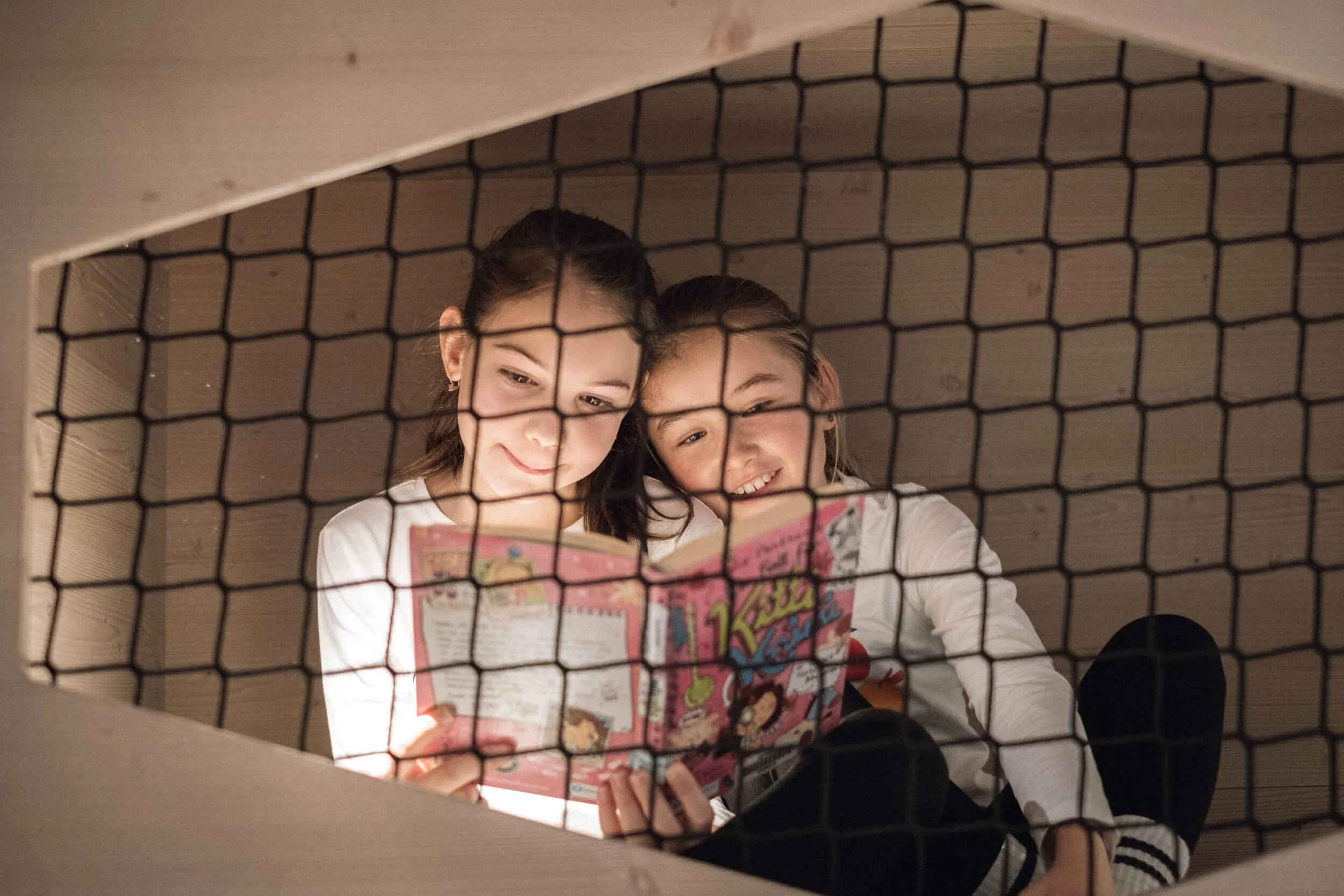 Zwei lachende Mädchen sitzen in einem Holzhaus, die durch ein Gitter betrachtet werden, während sie ein Kinderbuch lesen.