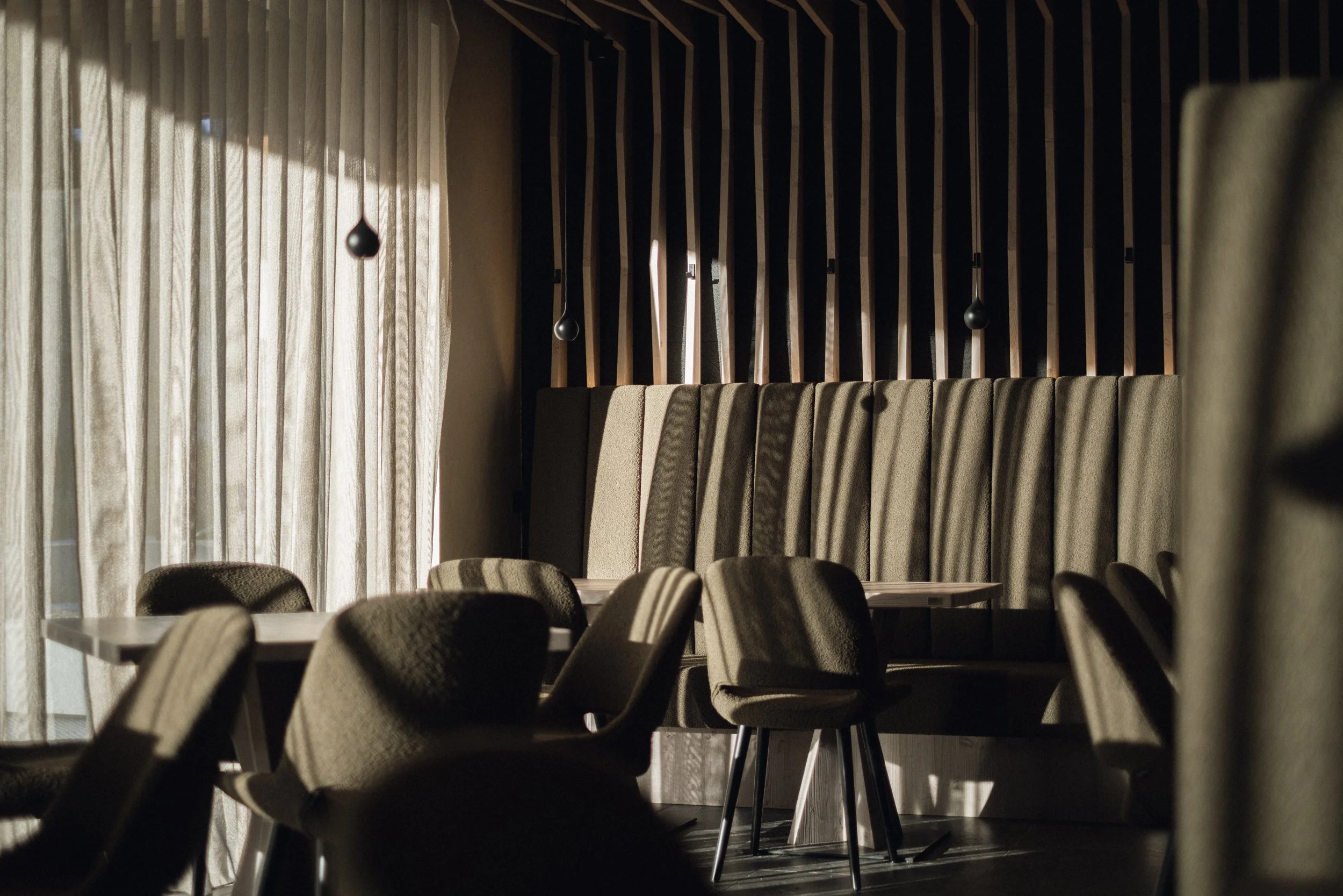Innenraum eines stilvollen, modernen Restaurants oder Cafés mit angehobenen Stühlen, Samt-Kopfstützen und dunklen Holzvertäfelung an der Wand, mit natürlichem Licht, das durch Vorhänge einfällt.