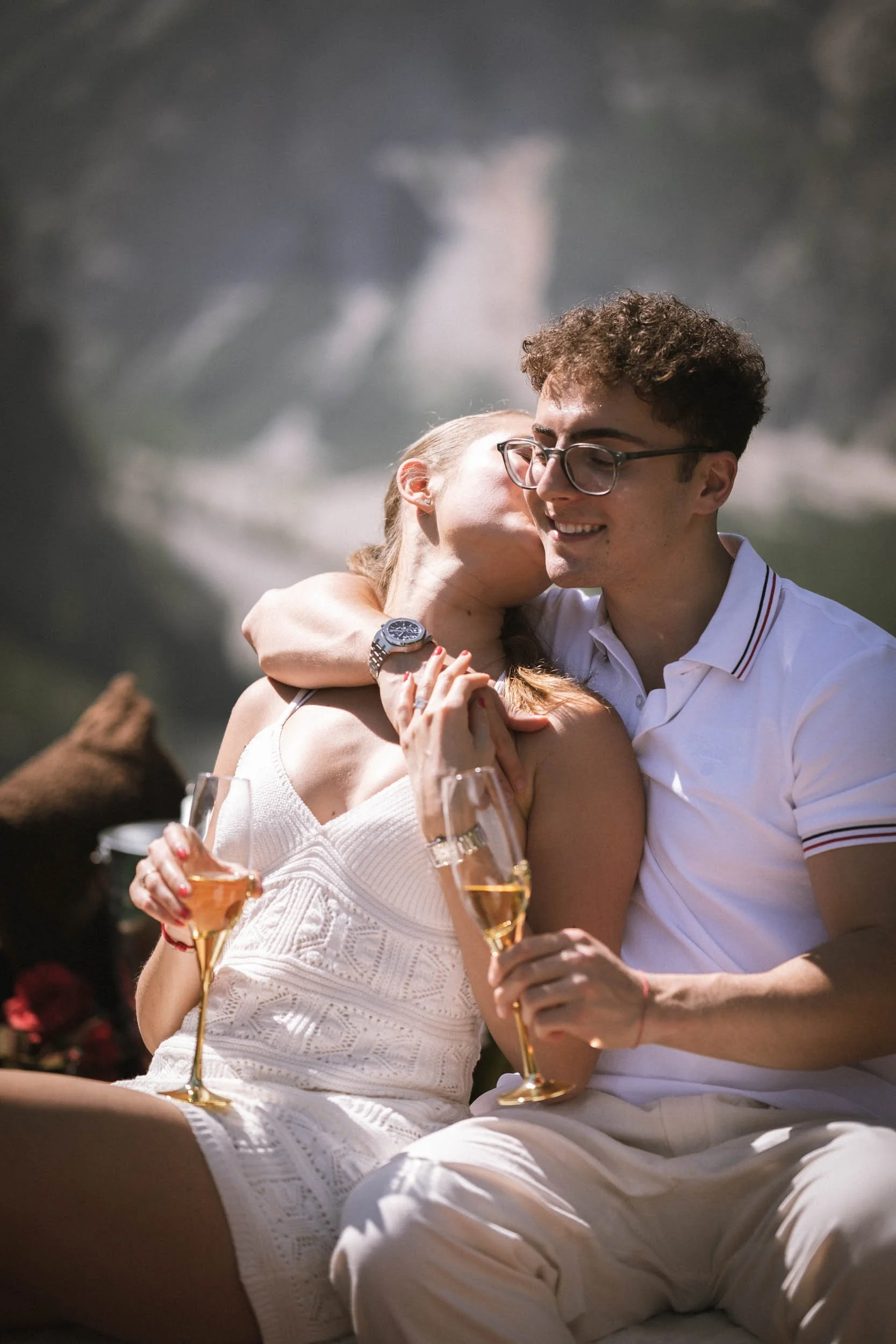 Ein junges Paar sitzt eng beieinander, trinkt Sekt und scheint sich küssen zu wollen, vor einer natürlichen Kulisse mit Wasserfall im Hintergrund.