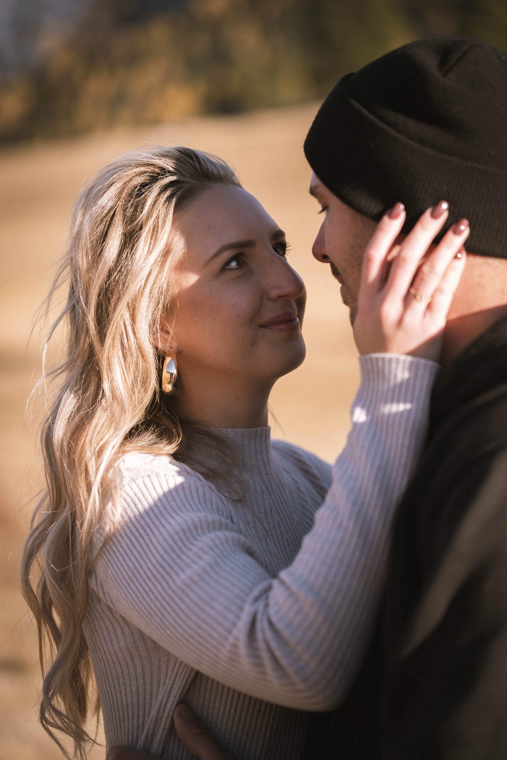 Ein Paar steht sich im Freien gegenüber, die Frau berührt die Nase des Mannes, während sie ihn liebevoll ansieht. Im Hintergrund befinden sich Bäume und eine goldene Herbstlandschaft.