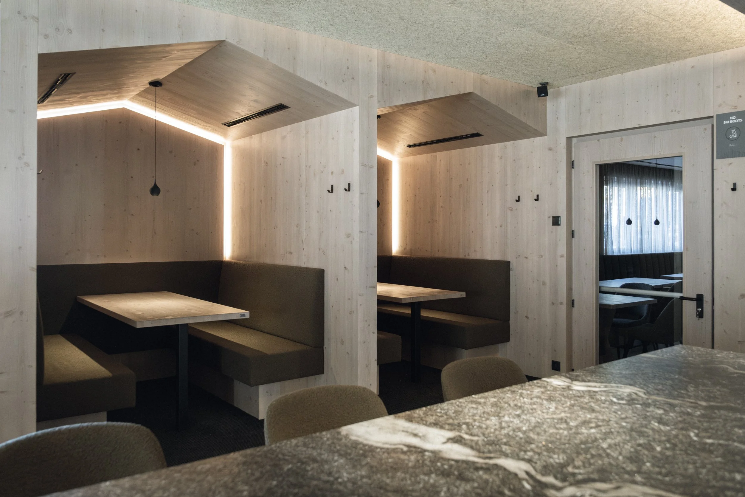 Innenansicht eines modernen, minimalistischen Restaurants mit Holzverkleidung, gepolsterten Sitzbänken, Tischen und einer separaten kleiner Kabine durch eine Tür mit Glas.