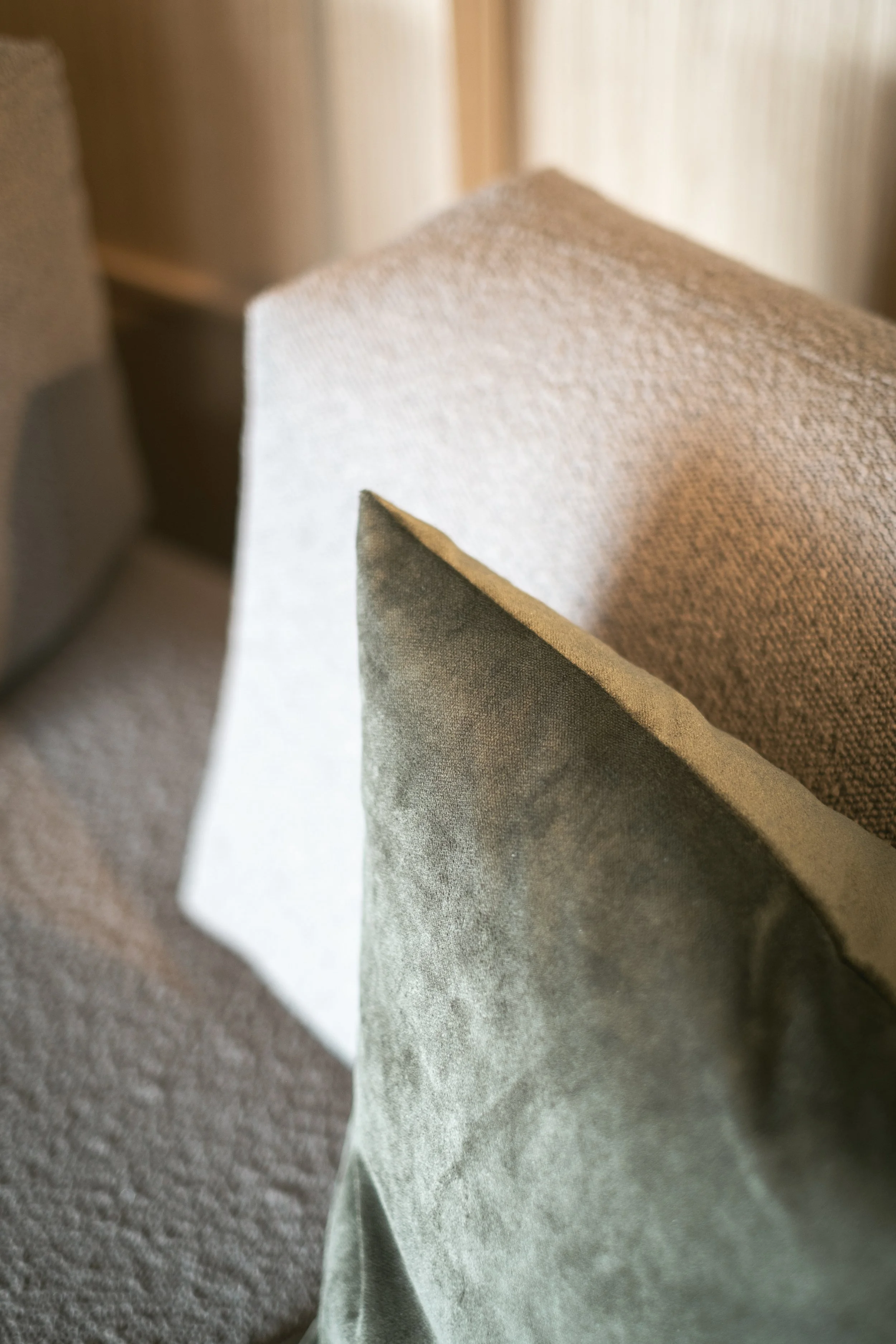 Nahaufnahme von drei verschiedenen Polstern in neutralen Tönen, auf einem Sofa oder Stuhl, mit Fokus auf die Stofftextur und das Ambiente eines Wohnzimmers.