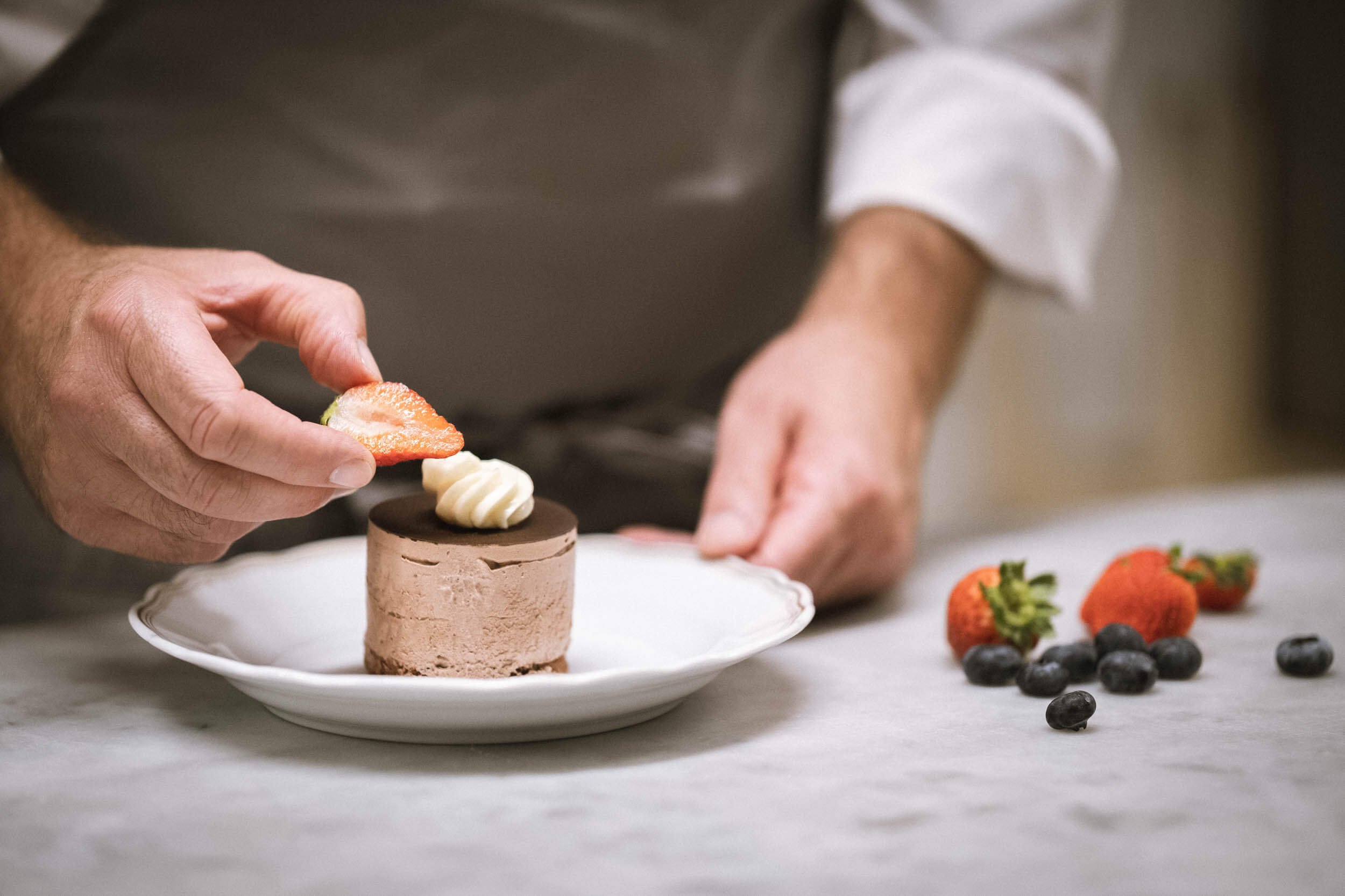 Nahaufnahme eines Köders, der auf einem weißen Teller platziert ist, während eine Person einen Erdbeerschnitz und einen Spritzgebäck auf einen Schokoladenmousse-Kuchen legt, mit Erdbeeren und Heidelbeeren auf einem weißen Tuch im Hintergrund.