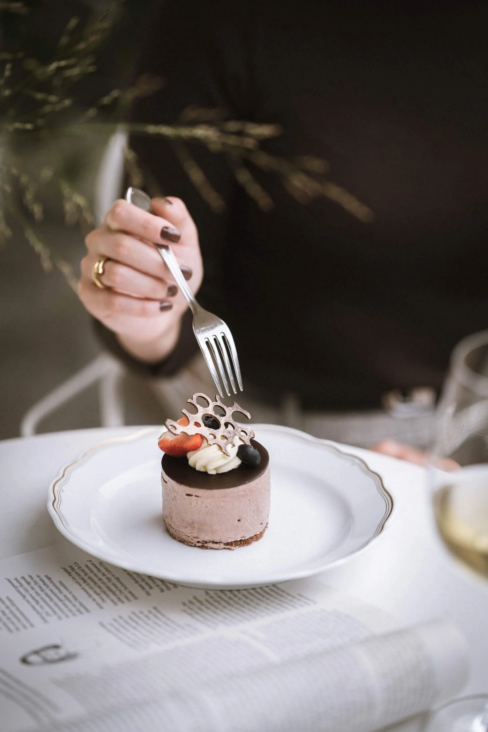 Eine Frau isst einen kleinen, runden Schokoladenkuchen auf einem weißen Teller, dekoriert mit Früchten und Schokoladenstückchen, während sie eine Gabel benutzt.