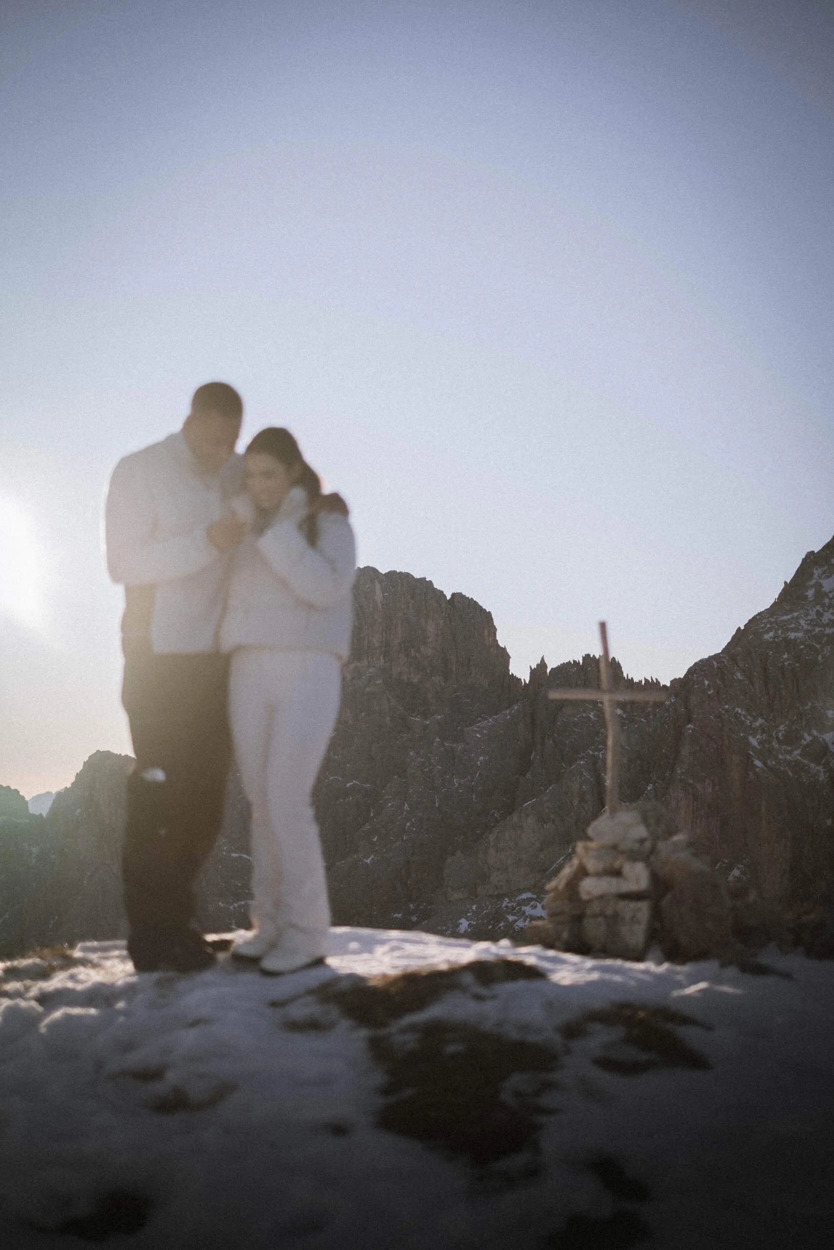 Ein Mann und eine Frau stehen gemeinsam auf einem schneebedeckten Berggipfel, umgeben von Bergen, mit einem kleinen Stein- und Holzkreuz im Vordergrund. Es ist Sonnenaufgang oder Sonnenuntergang im Hintergrund.