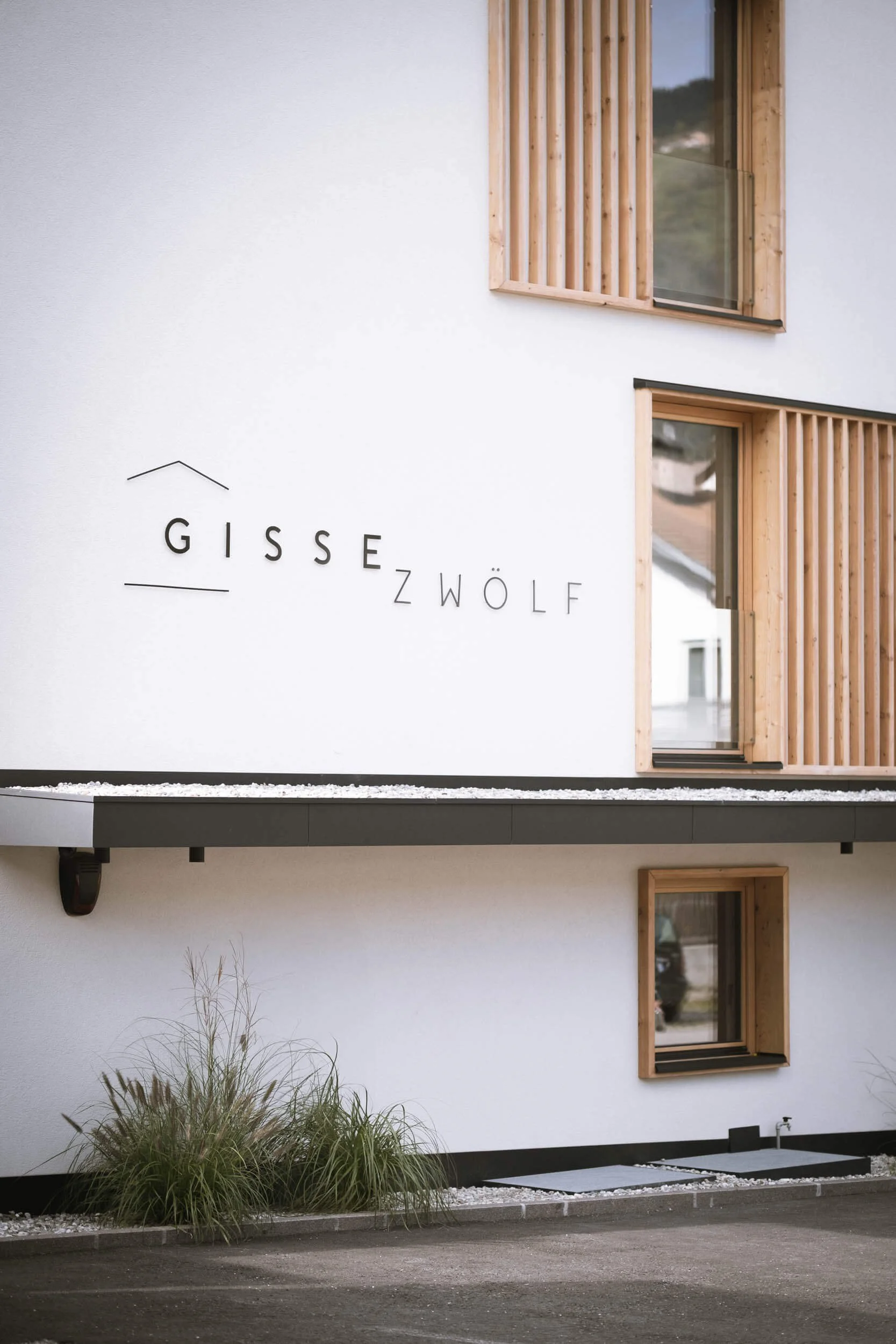 Frontansicht eines weißen Gebäudes mit Holzfenstern und Holzläden, die an einer modernen Fassade angebracht sind. Das Gebäude hat die Beschriftung 'GISS Z WÖLF' an der Wand.