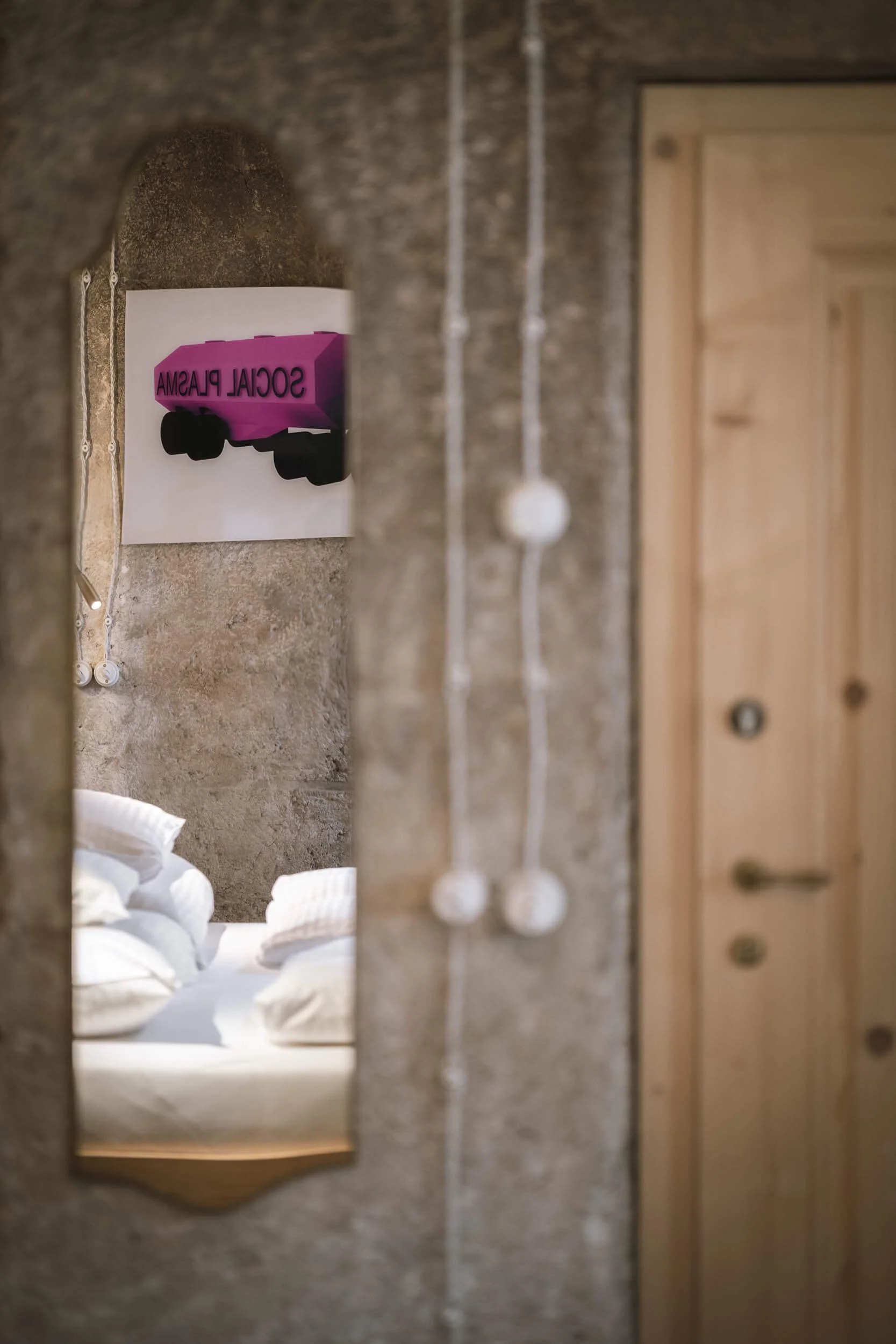 Spiegel in einem rustikalen Raum, der eine Ecke eines Bettes mit weißen Bettwaren zeigt, eine Wand mit einem Poster und einer Lampe mit weißen Kugeln. Eine Holztür ist sichtbar.