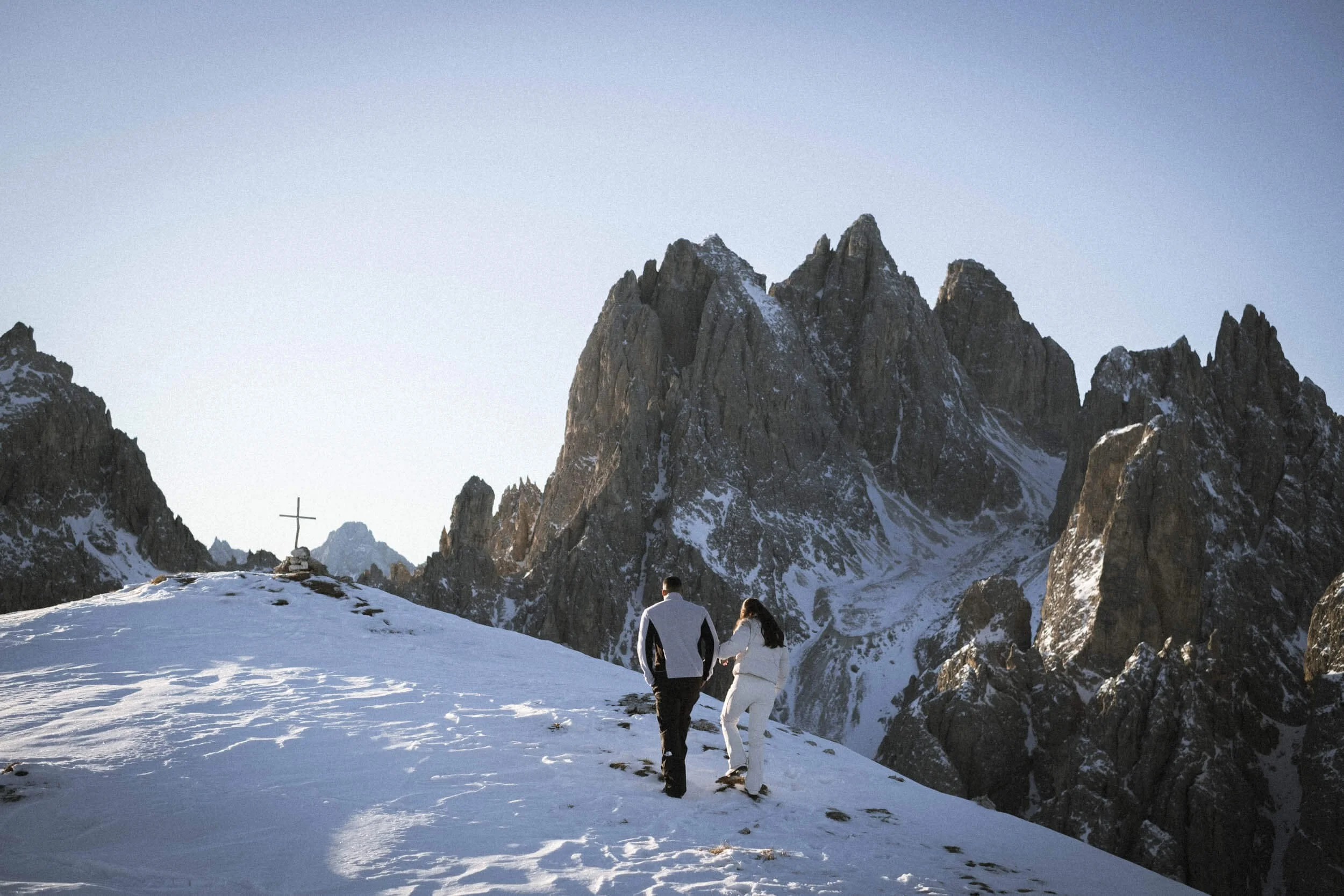 Ein Paar geht Hand in Hand auf schneebedecktem Bergpfad mit hohen, schroffen Bergen im Hintergrund, ein Kreuz auf einem kleinen Hügel am Horizont.