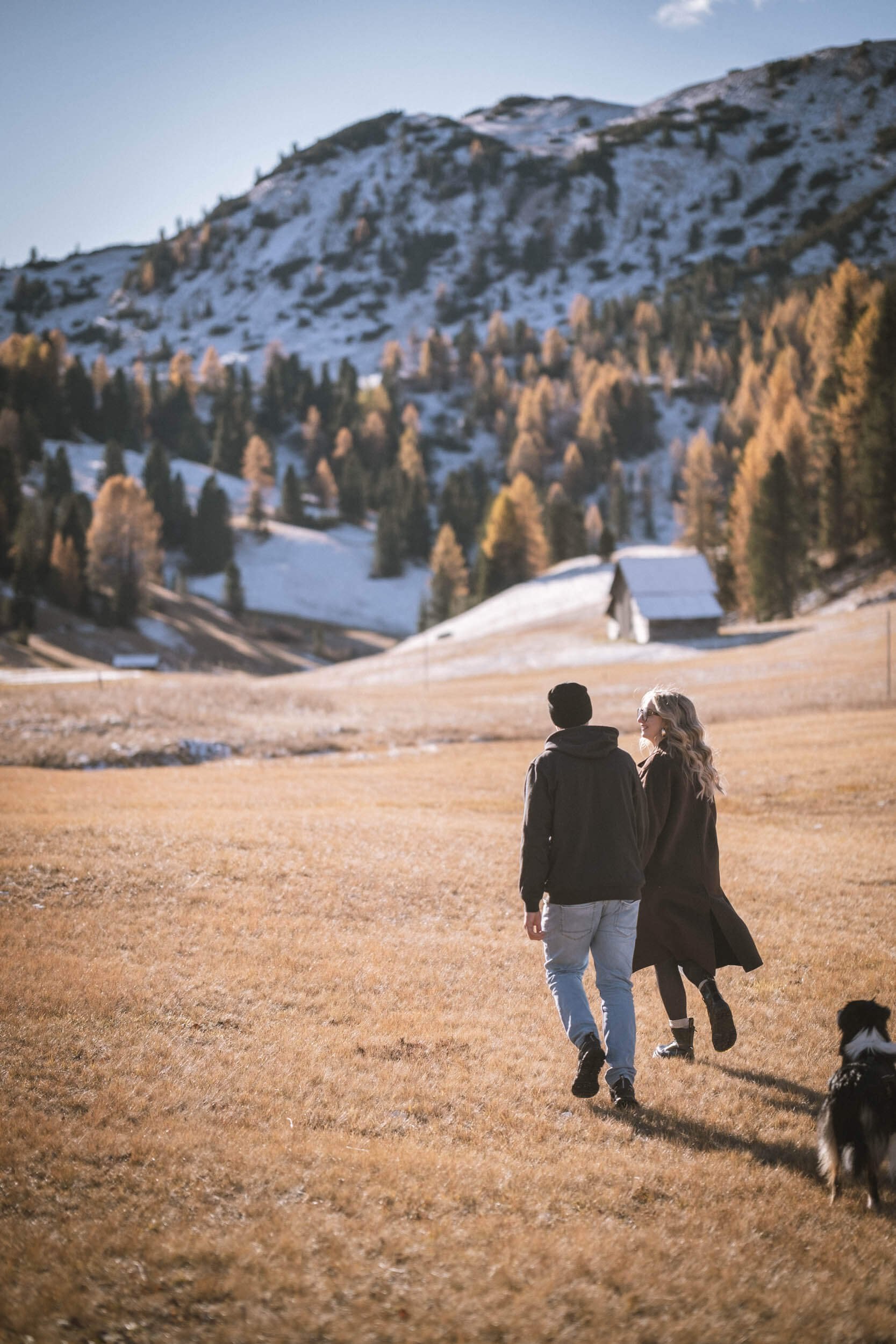 Ein Mann und eine Frau laufen auf einer weiten Wiese in einer Bergregion, begleitet von einem Hund.