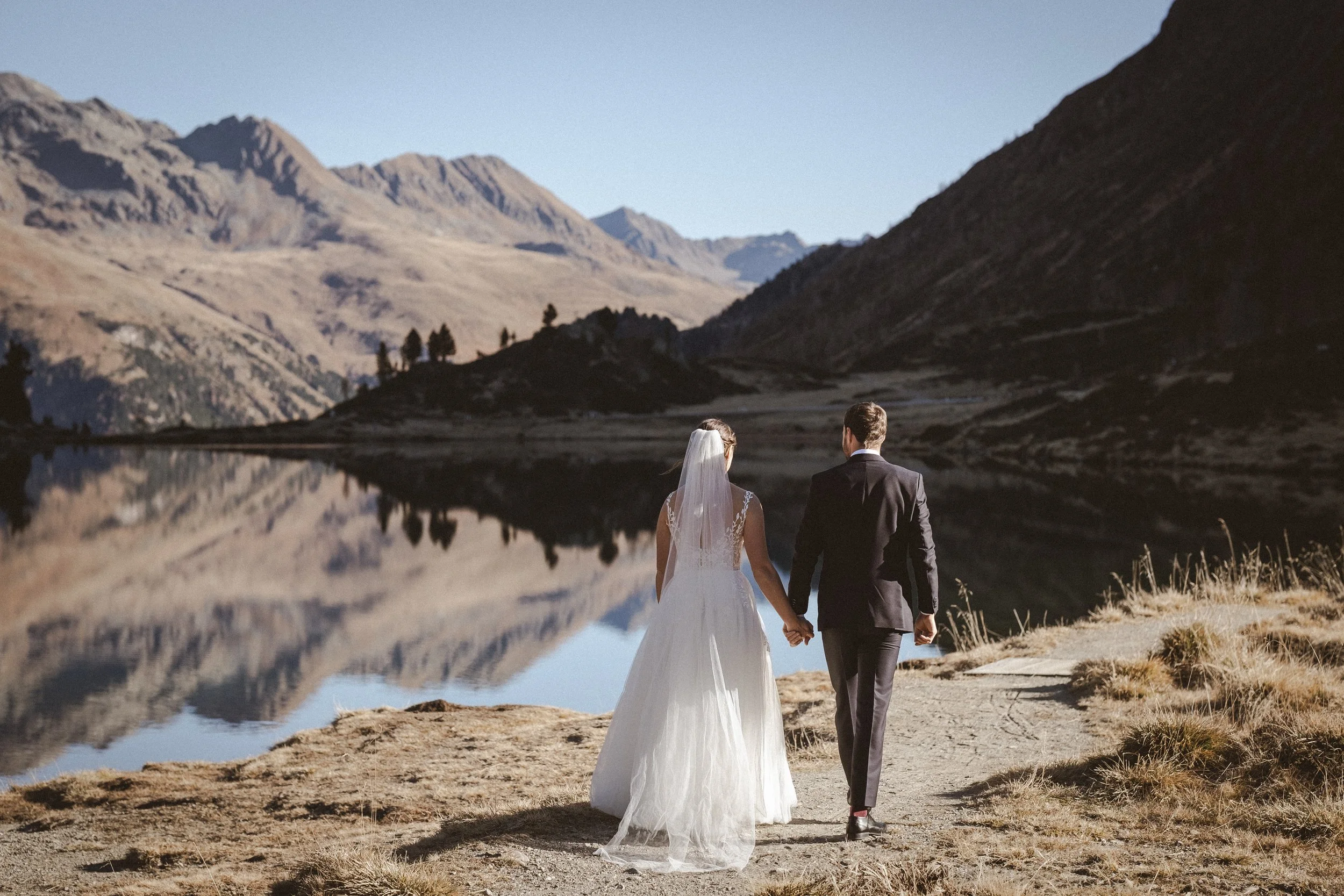 Ein Brautpaar hält Händchen am See mit Bergen im Hintergrund.