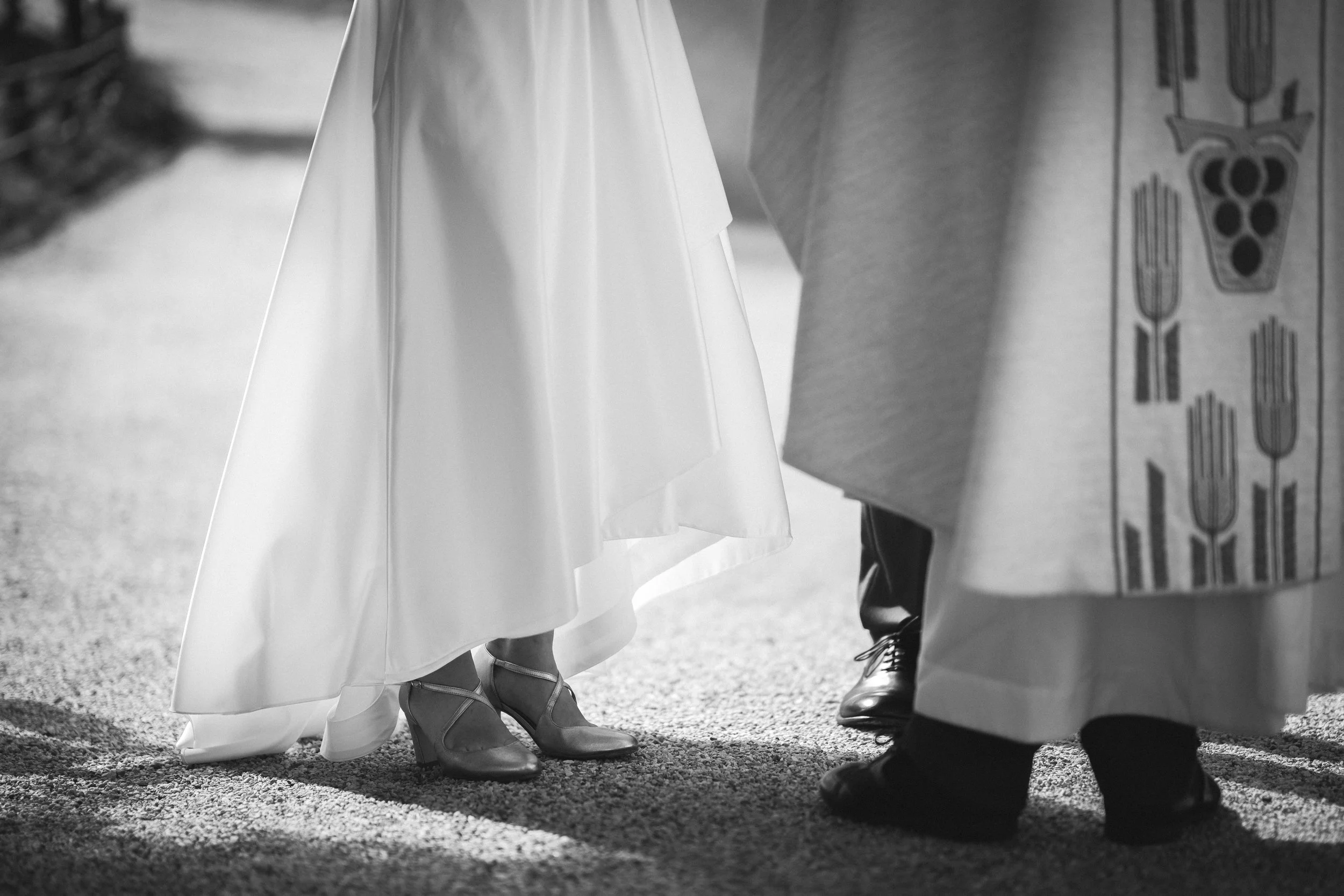Nahaufnahme von einem Brautpaar, das sich bei einer Hochzeitgegenübersteht, mit Fokus auf die Schuhe und den unteren Kleidungsbereich, im Schwarz-Weiß-Format.