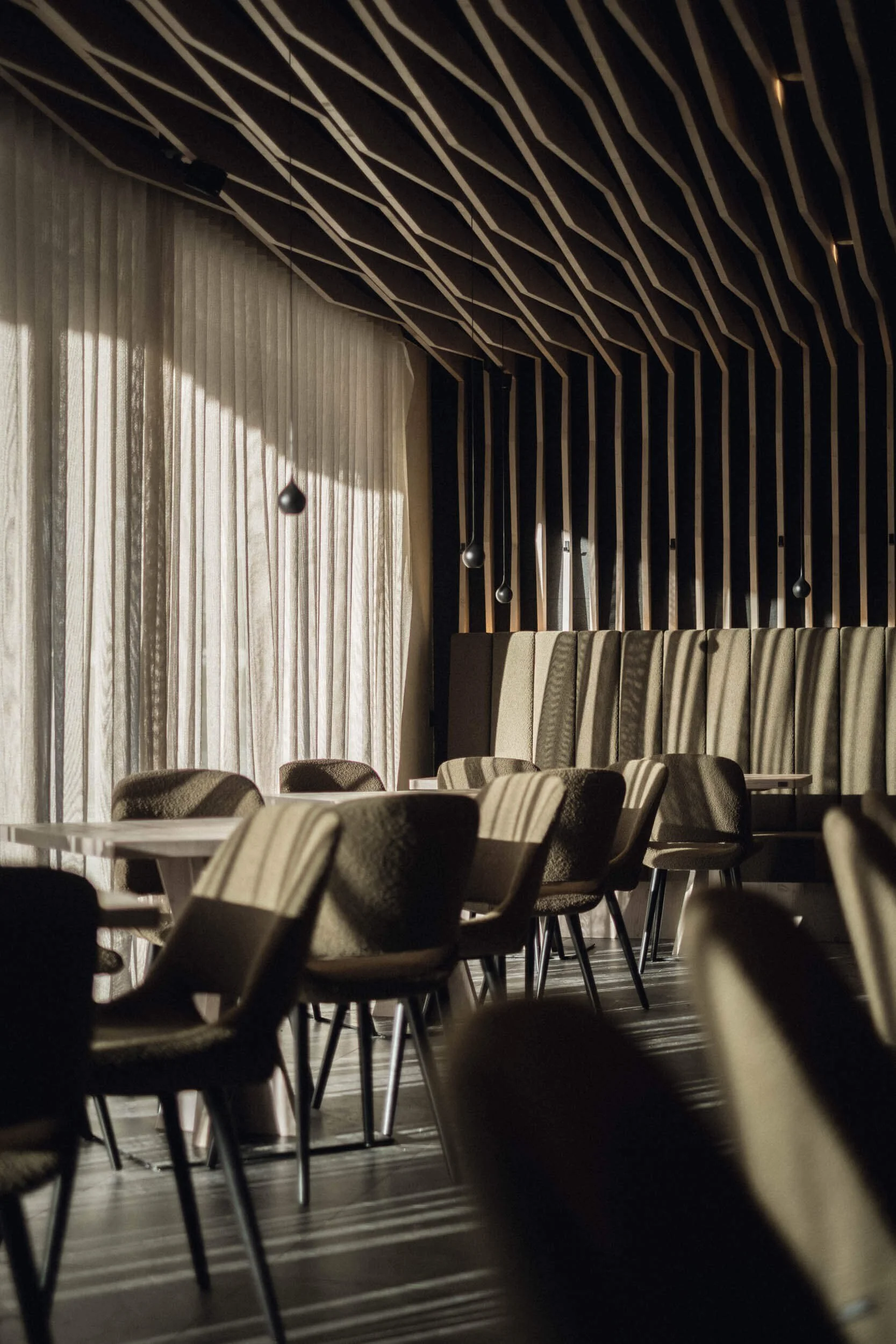 Leerer Speisesaal mit Stühlen und Tischen, Sonnenlicht durch Fenster, dunkle Holzdecke mit gestreiftem Design, elegante Vorhänge.