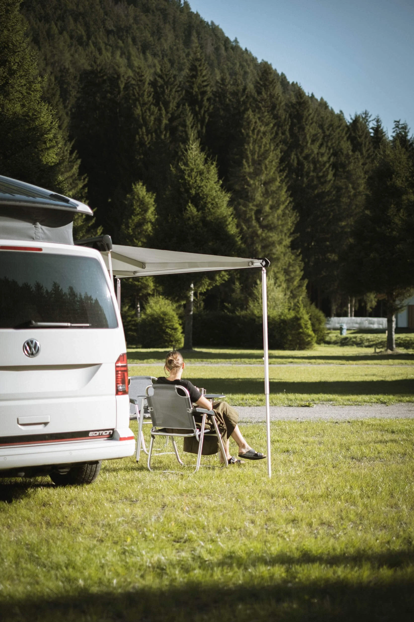 Person sitzt in einem Klappstuhl vor einem weißen Wohnmobil im Freien, mit Blick auf einen Wald und eine grüne Wiese, bei sonnigem Wetter.