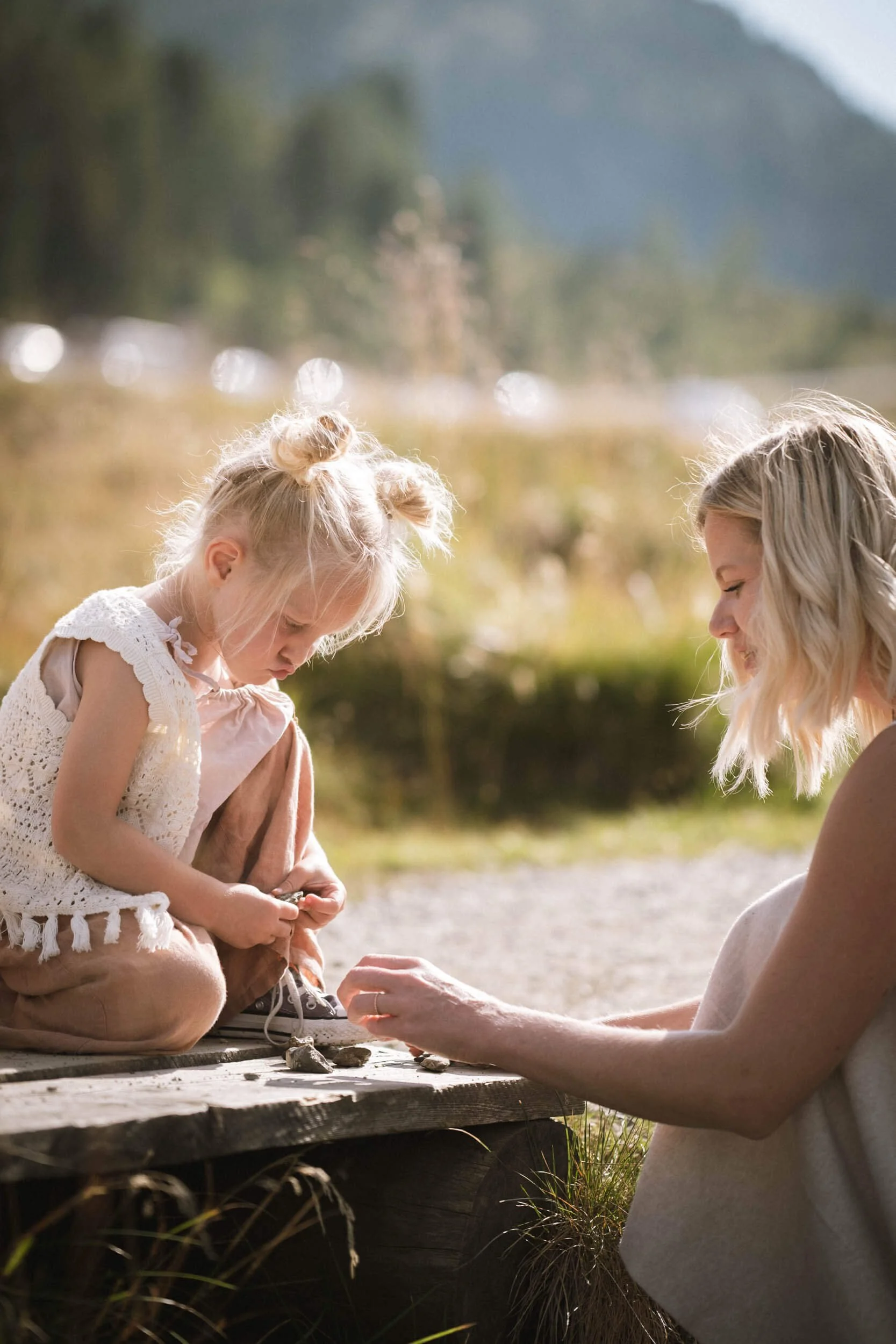 Eine junge Frau sitzt im Grünen und stellt eine kleine Mädchen auf ein Holzstamm, während sie gemeinsam Steine sammeln.