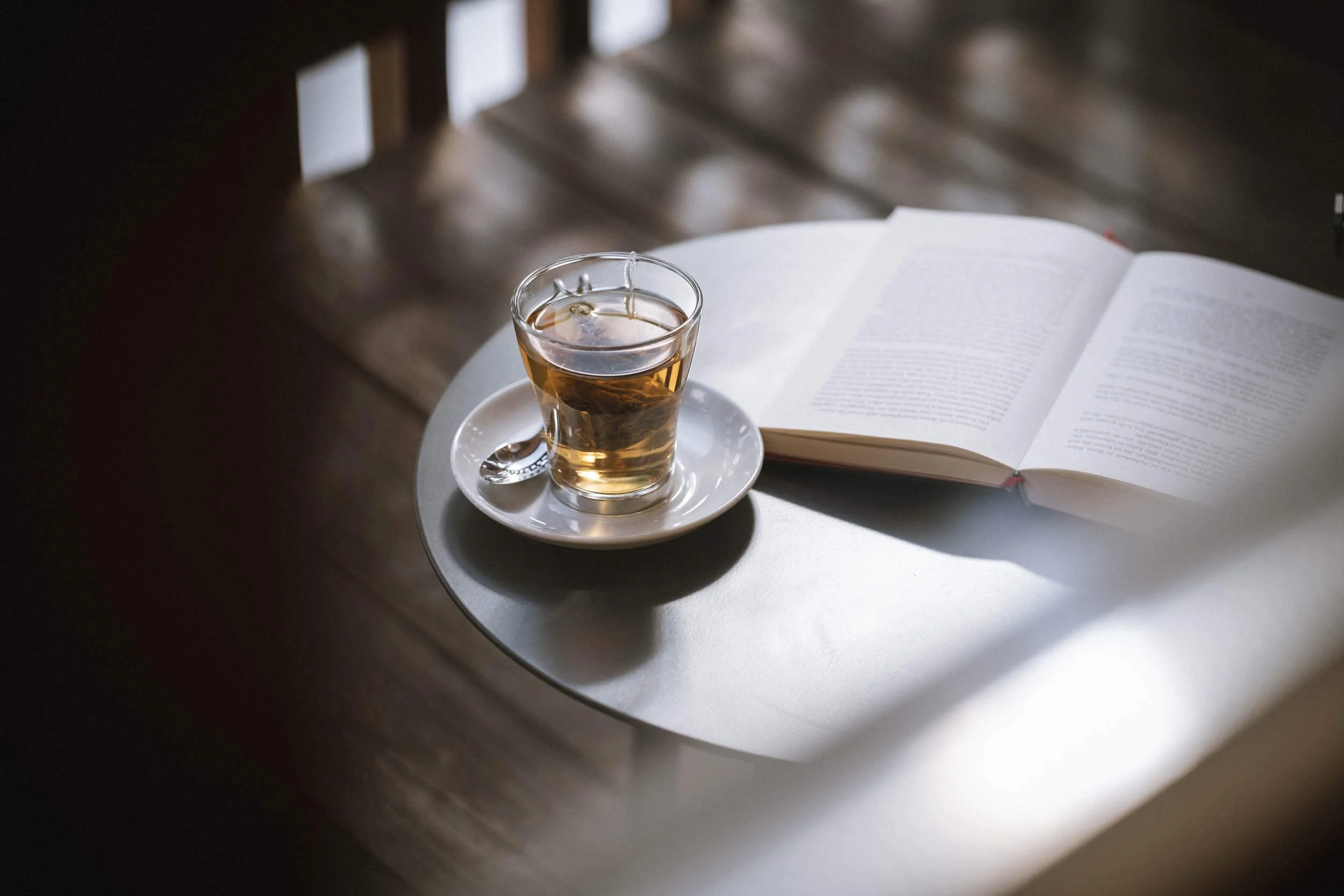 Eine Tasse Tee auf einem weißen Unterteller mit Löffel neben einem offenen Buch auf einem runden weißen Tisch bei natürlichem Licht.