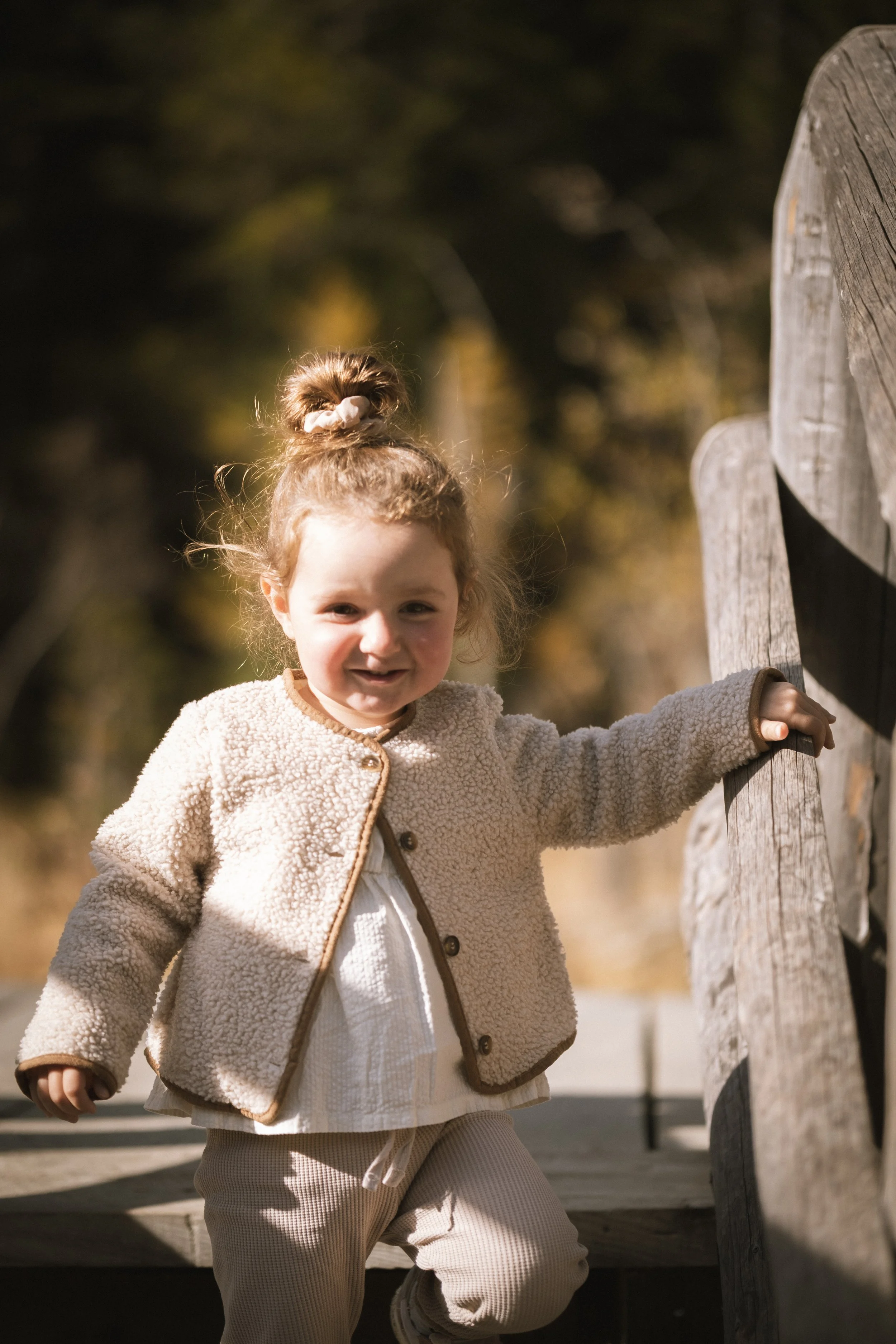 Ein kleines Mädchen mit lockigem Haar in einem hohen Pferdehaarlost, beige Jacke und hellen Hosen, das an einem Holzgeländer steht und in die Kamera lächelt.