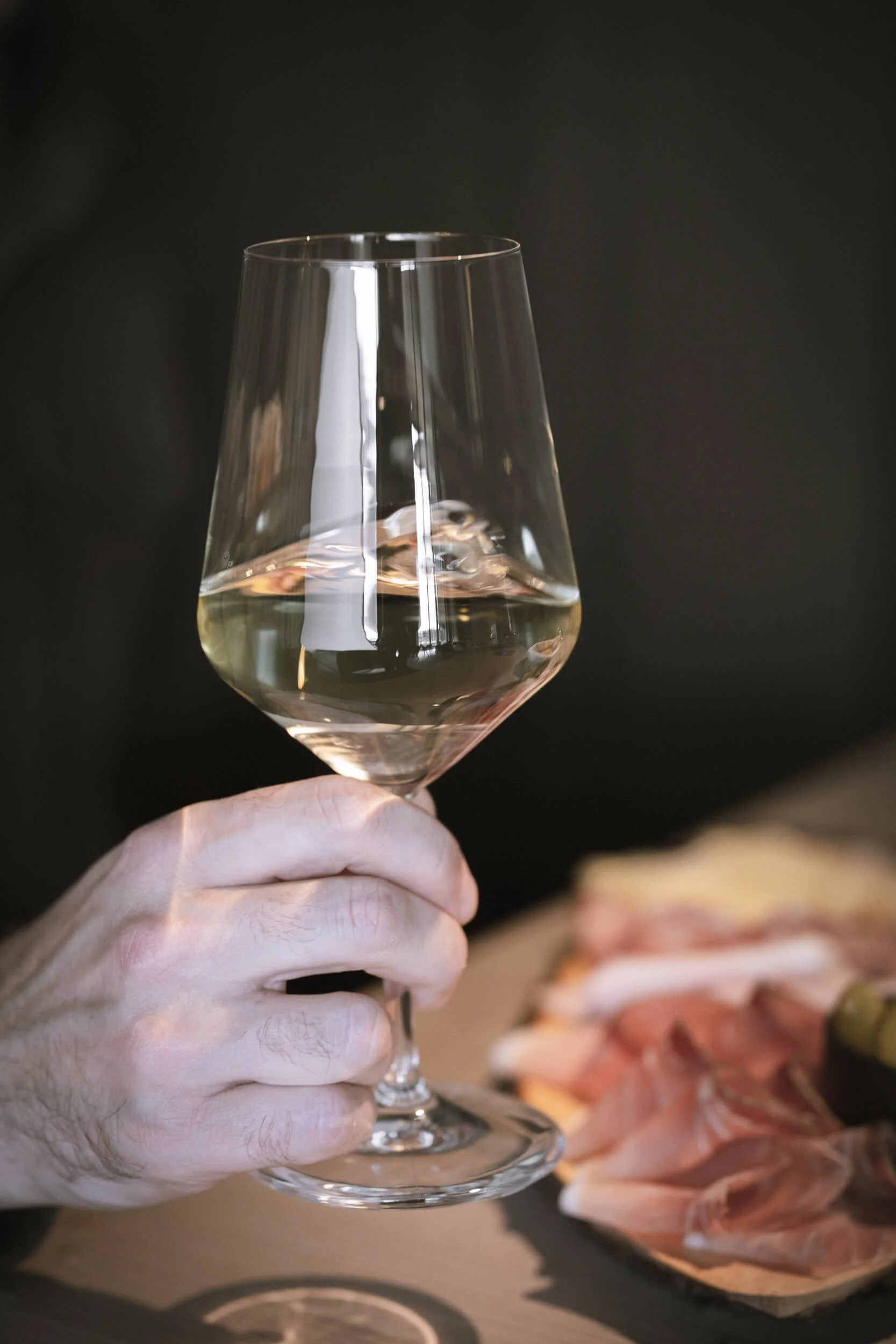 Person hält ein Weinglas mit weißem Wein, im Hintergrund eine Platte mit Aufschnitt. Dunkler Hintergrund.