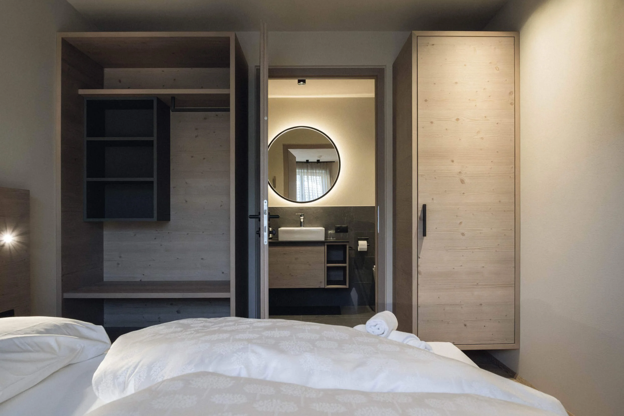 Schlafzimmer mit offener Tür zum Badezimmer, Doppelbett mit weißen Kissen und Handtüchern, moderne Holzschränke und Wanddeko, runder Spiegel im Bad, Lichtreflex auf Bett