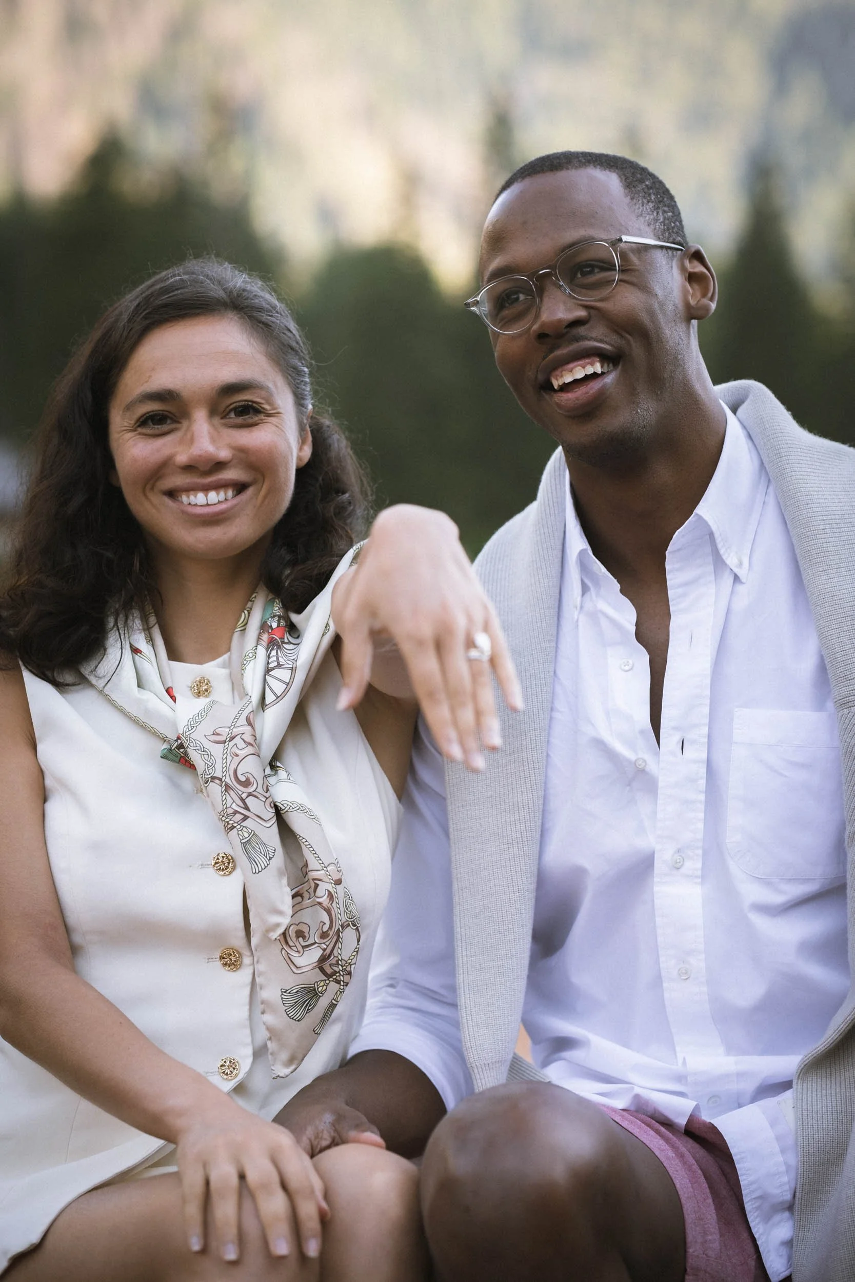 Ein lachendes weißes Paar sitzt im Freien, umgeben von Natur, beim Treffen oder Treffen im Park, mit einem entspannten, fröhlichen Ausdruck.