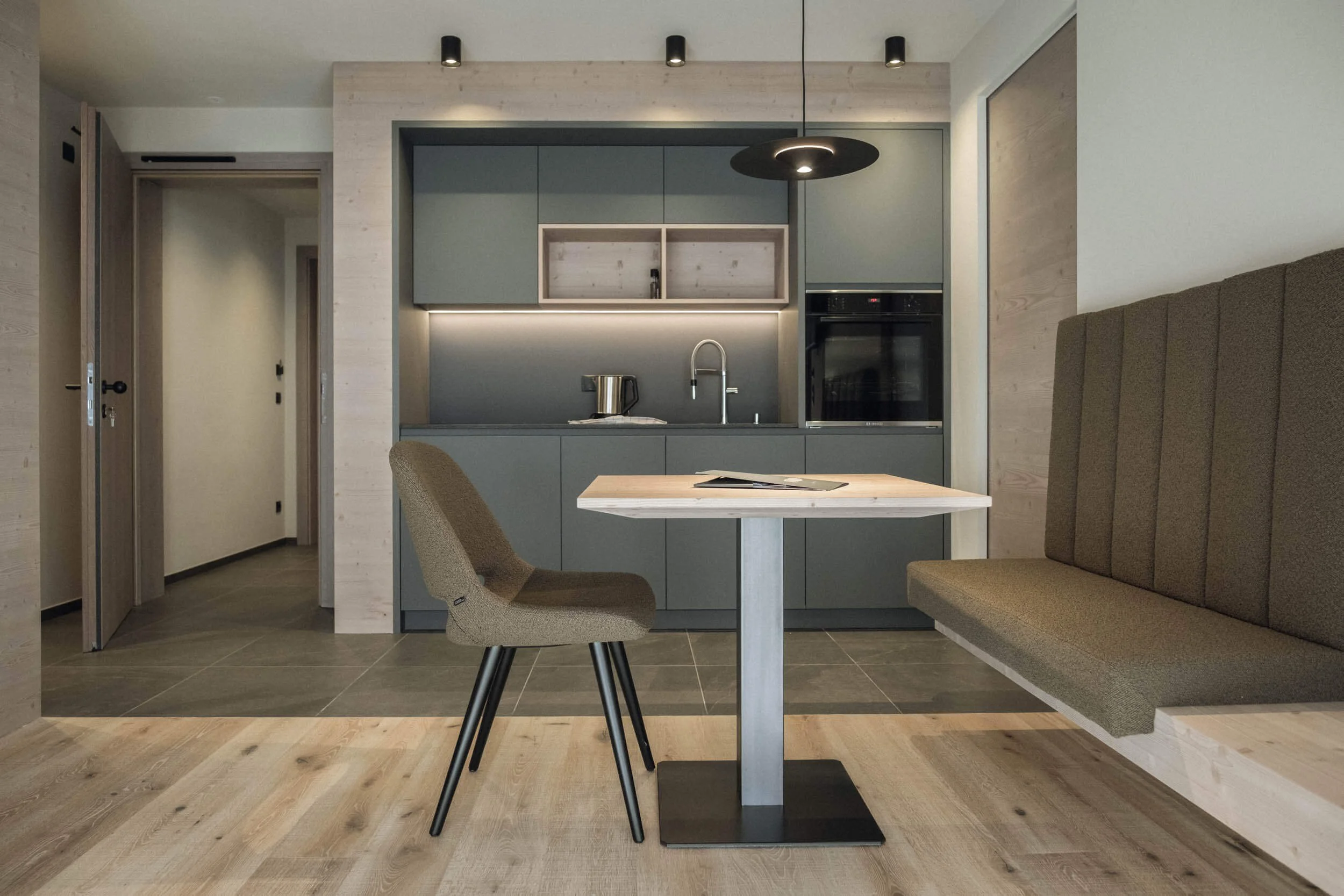 Moderne minimalistische Küche mit grau Schränken, grauem Esstisch, grauem Stuhl und einer Sitzbank, heller Holzboden, dunkler Fliesenspiegel, Esszimmer in einem Apartment.
