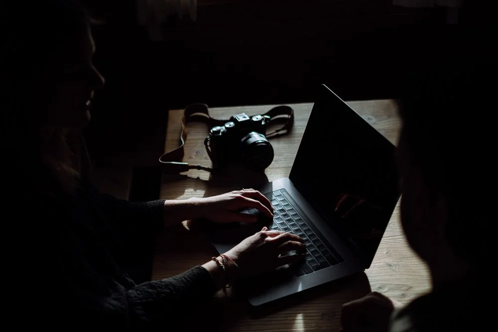 Zwei Personen arbeiten an einem dunklen Laptop auf einem Holztisch, mit einer Kamera im Hintergrund.