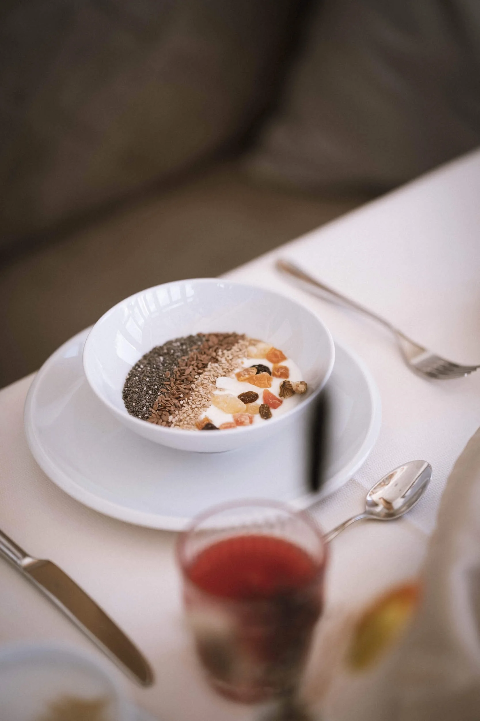 Schale mit Joghurt und verschiedenen Samen und Trockenfrüchten, auf einem weißen Teller mit Besteck daneben.