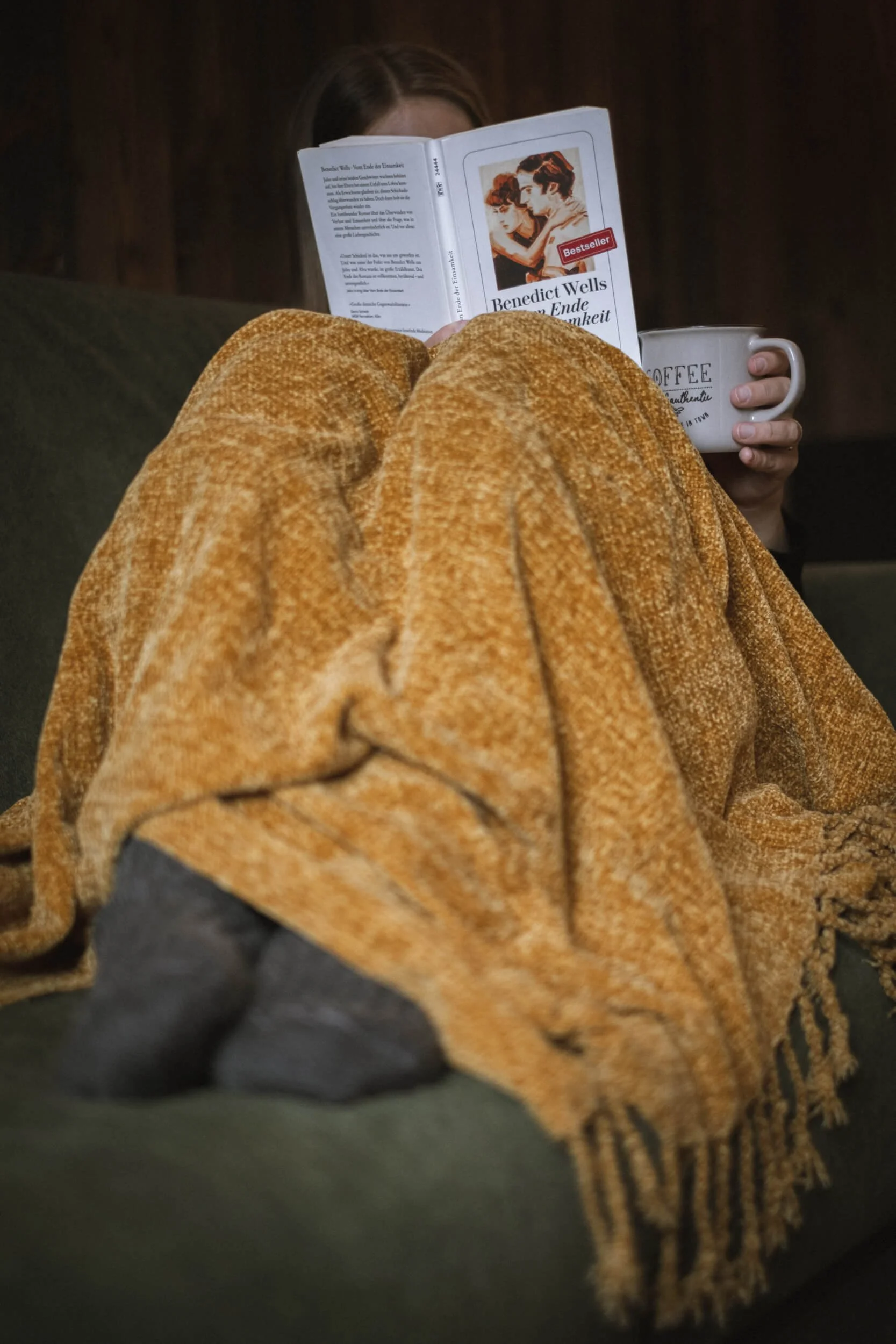 Person liest ein Buch und hält eine Tasse, während sie es sich auf einem Sofa gemütlich macht, zugedeckt mit einer gelben Decke.