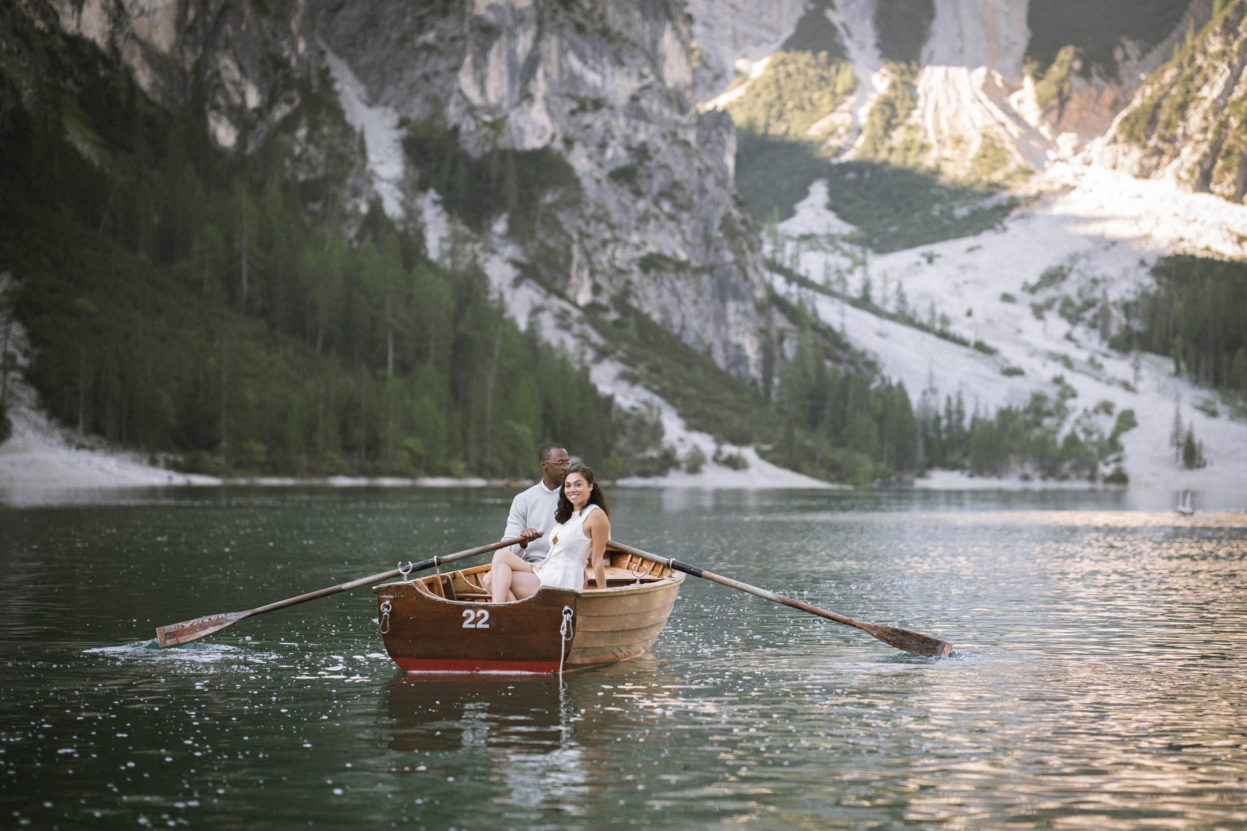 Ein Paar sitzt in einem kleinen Holzboot auf einem See inmitten einer bergigen Landschaft mit Wäldern und weißen Felsen im Hintergrund.