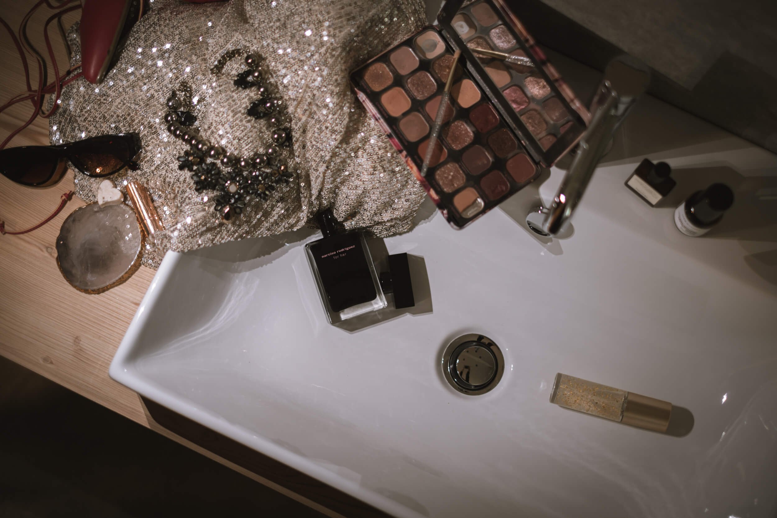 Wäscheschale mit Make-up-Produkten, Parfüm, Sonnenbrille, Schmuck, Nagellack, Kosmetiktücher, und weitere Kosmetika auf einem Waschtisch.