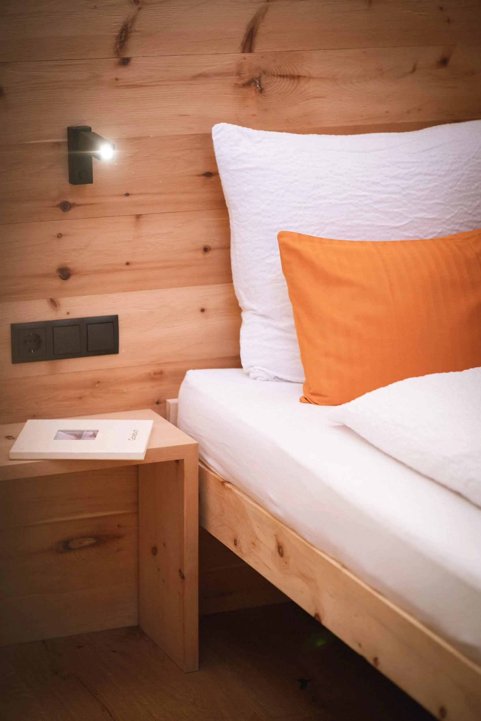 Nahaufnahme eines Bettes mit weißer Bettwäsche, einer orangefarbenen Kissenhülle, einem Holz-Nachttisch und einer Wandlampe an einer Wand aus Holzpaneelen.