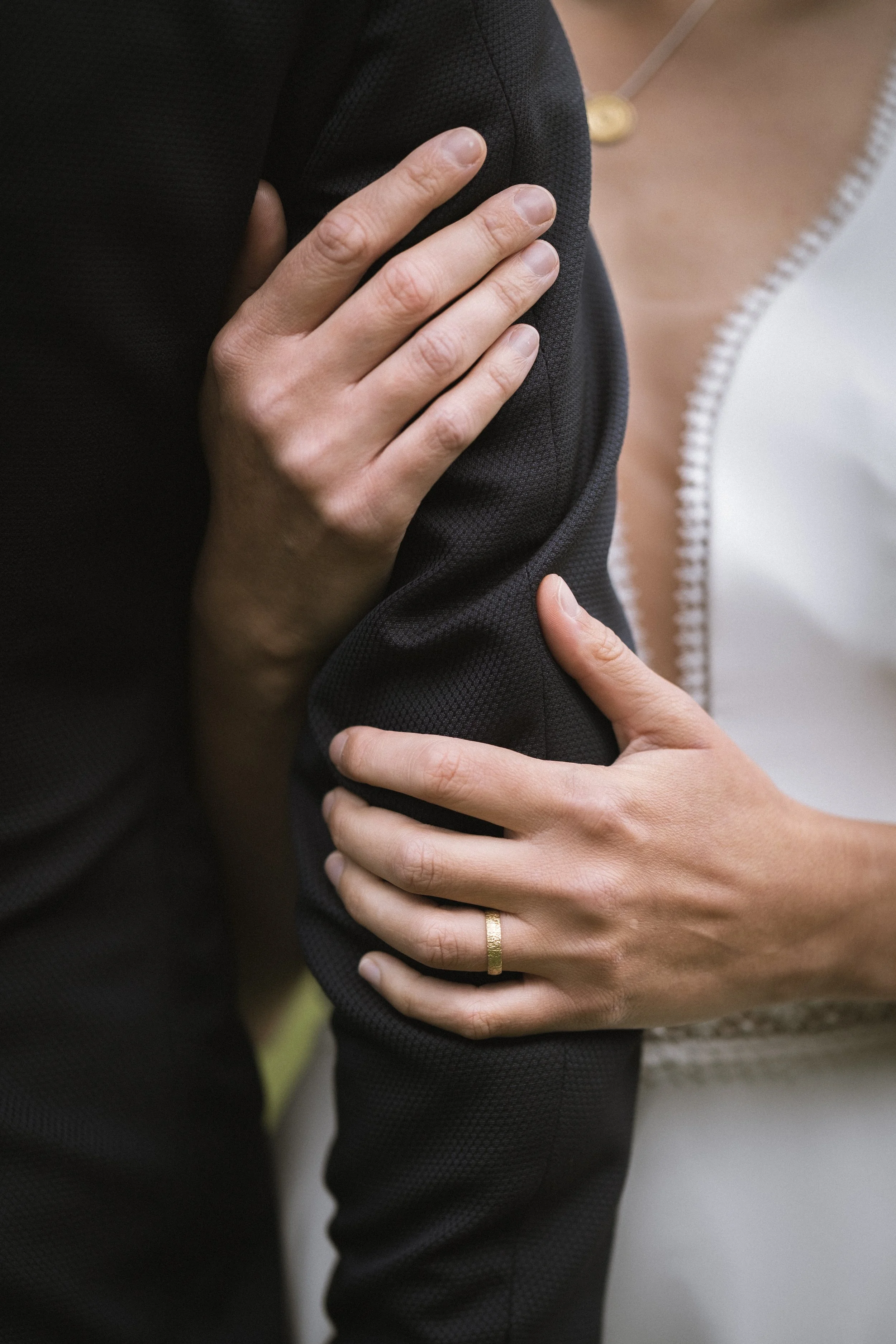 Nahaufnahme von zwei Händen, die zärtlich einander am Oberarm und Hand halten, bei einer Person in einem schwarzen Outfit und einer Frau mit einem Ring an der Hand.