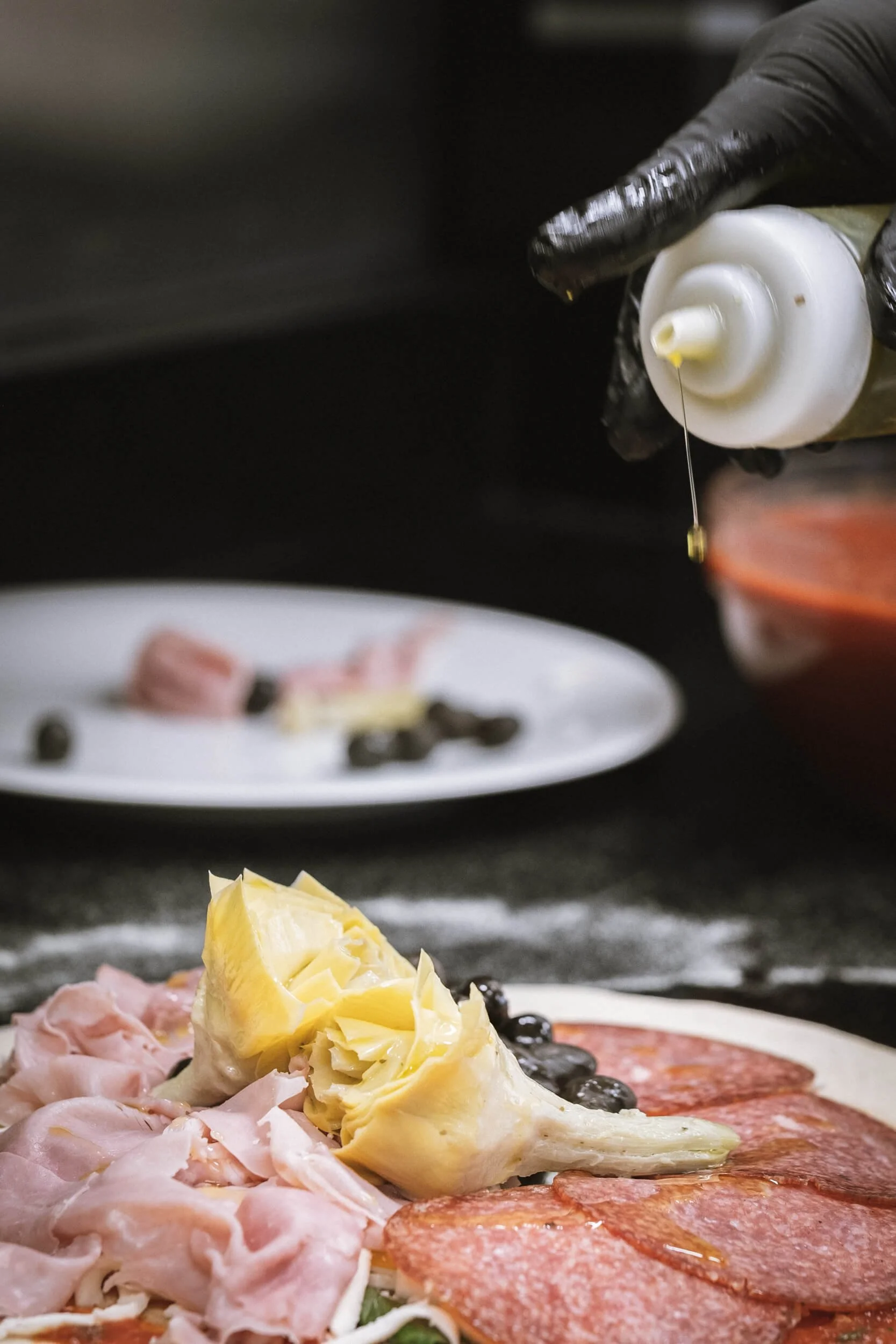 Auf einer schwarzen Oberfläche wird eine Pizza mit Käse, Schinken und Salami zubereitet. Eine Hand, die eine Flasche mit Öl oder Tomatensauce hält, wird darüber gehalten.