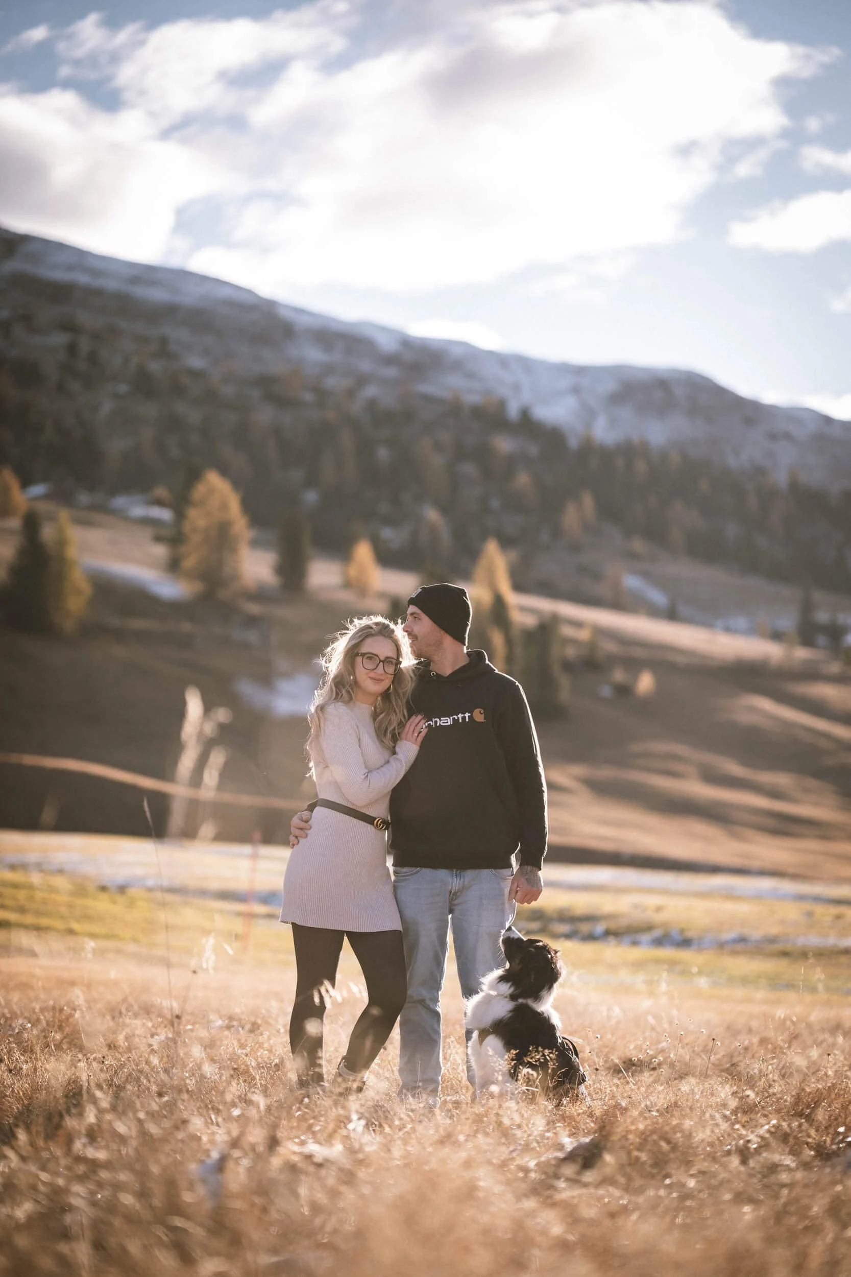 Ein Paar mit Hund in einer ländlichen, bergigen Landschaft bei Sonnenuntergang.
