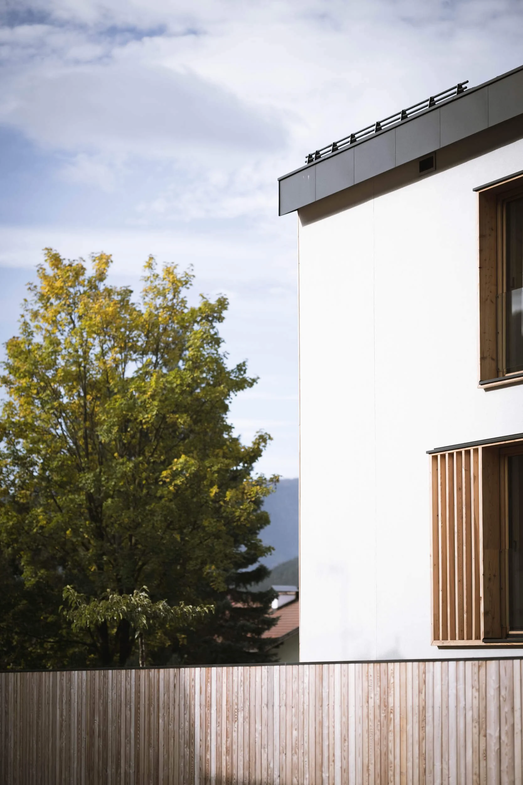 Moderne weiße Hausfassade mit Holzfenstern und Zaun, Baum im Vordergrund, bewölkter Himmel im Hintergrund.