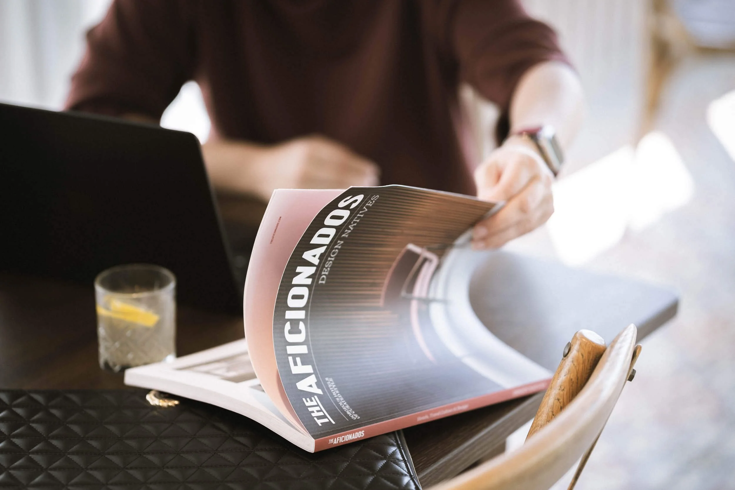 Ein aufgeschlagenes Magazin namens 'The Afficionado's' liegt auf einem Tisch, neben einem Trinkglas mit Wasser und Zitronenscheibe. Im Hintergrund ist eine Person, die das Magazin liest.