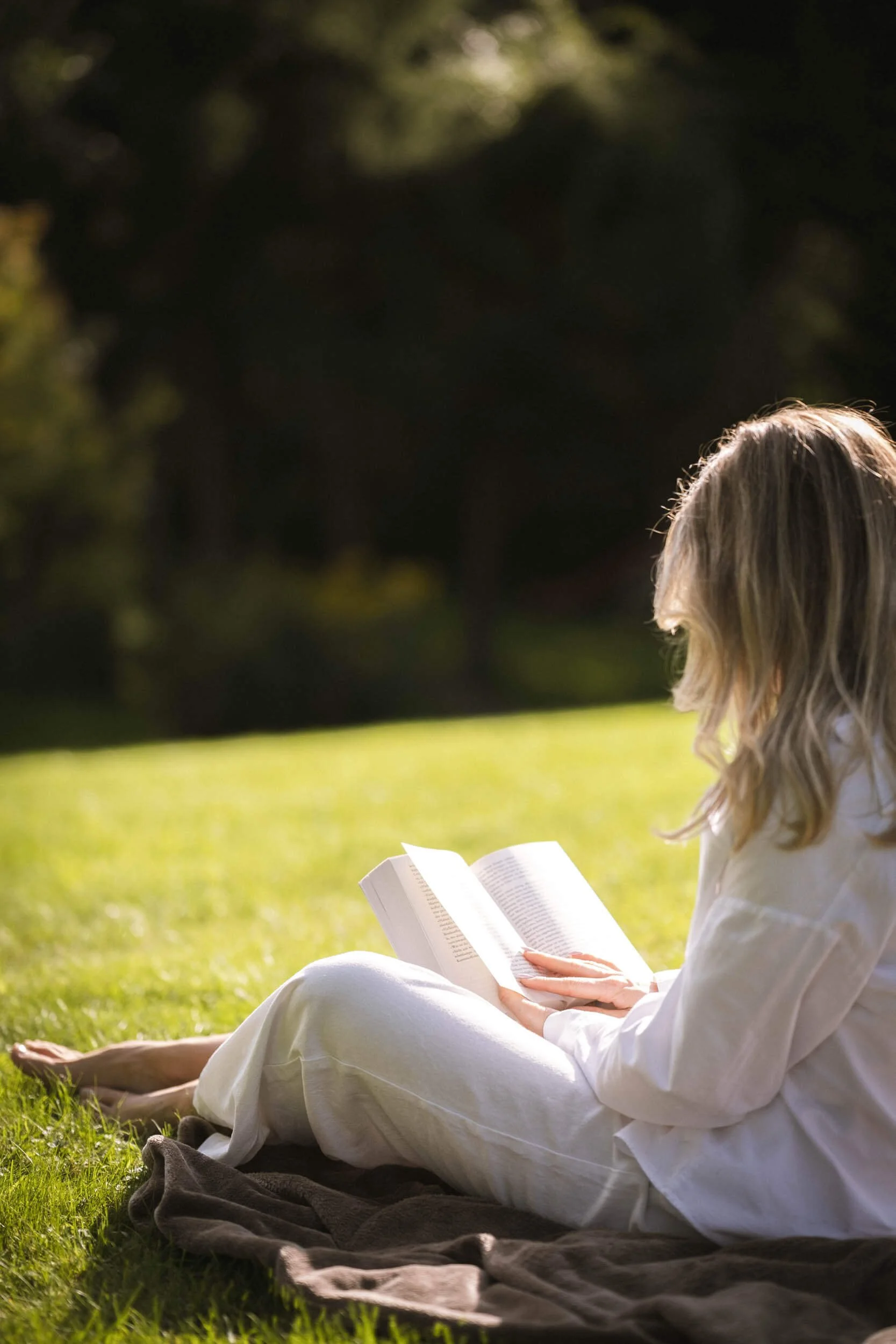 Frau liest entspannt im Freien auf einer Wiese, sitzend auf einer Decke, bei Sonnenlicht, mit Bäumen im Hintergrund.