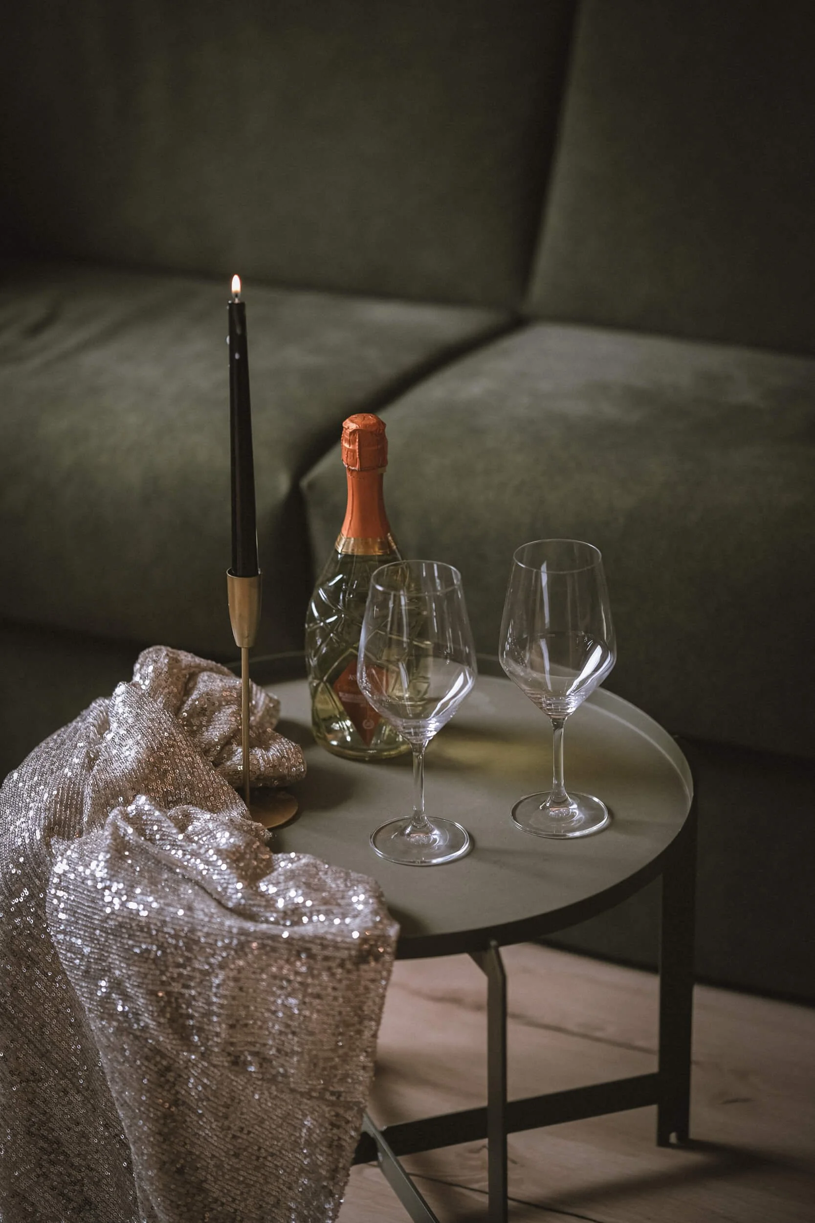 Ein Tisch mit einer Weinflasche, zwei leeren Weingläser, einer Kerze und einem silbernen Stoff, vor einem grauen Sofa.