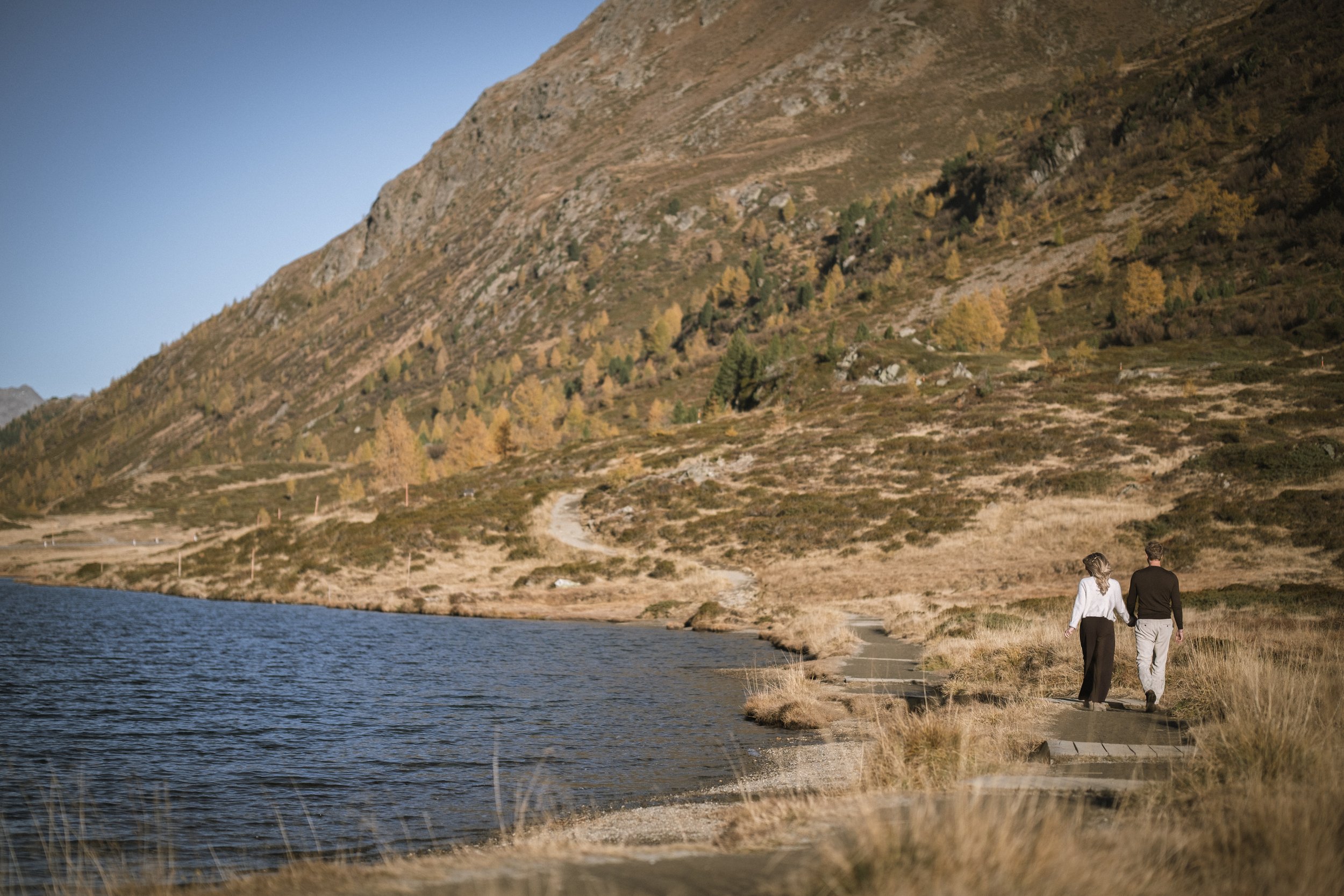 Ein Paar spaziert Hand in Hand an einem See in einer bergigen Landschaft im Herbst.