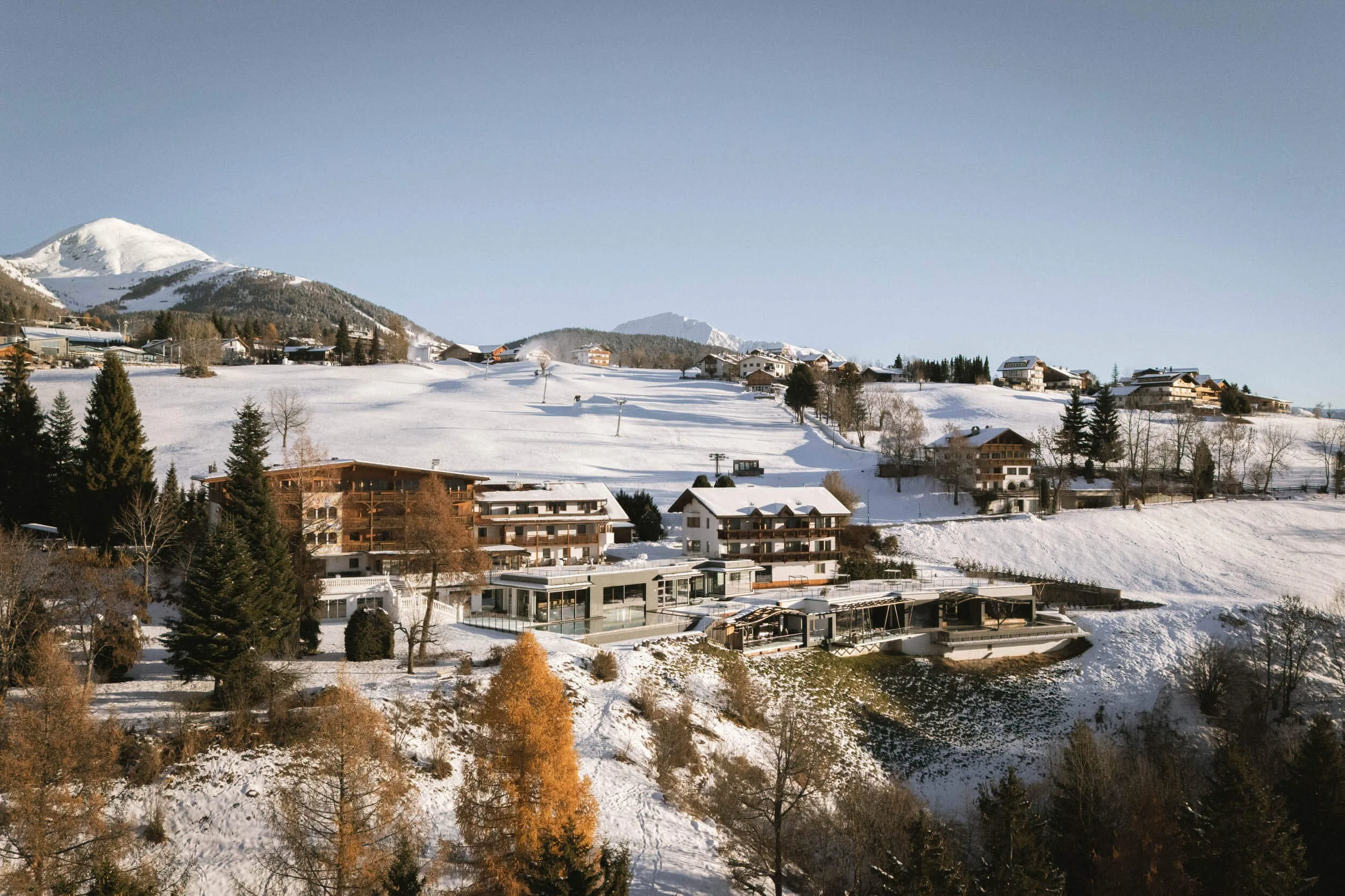 Ein verschneiter Bergort mit modernen Gebäuden im Vordergrund, umgeben von Bäumen und offenen Schneeflächen, mit Bergen im Hintergrund bei Sonnenlicht.