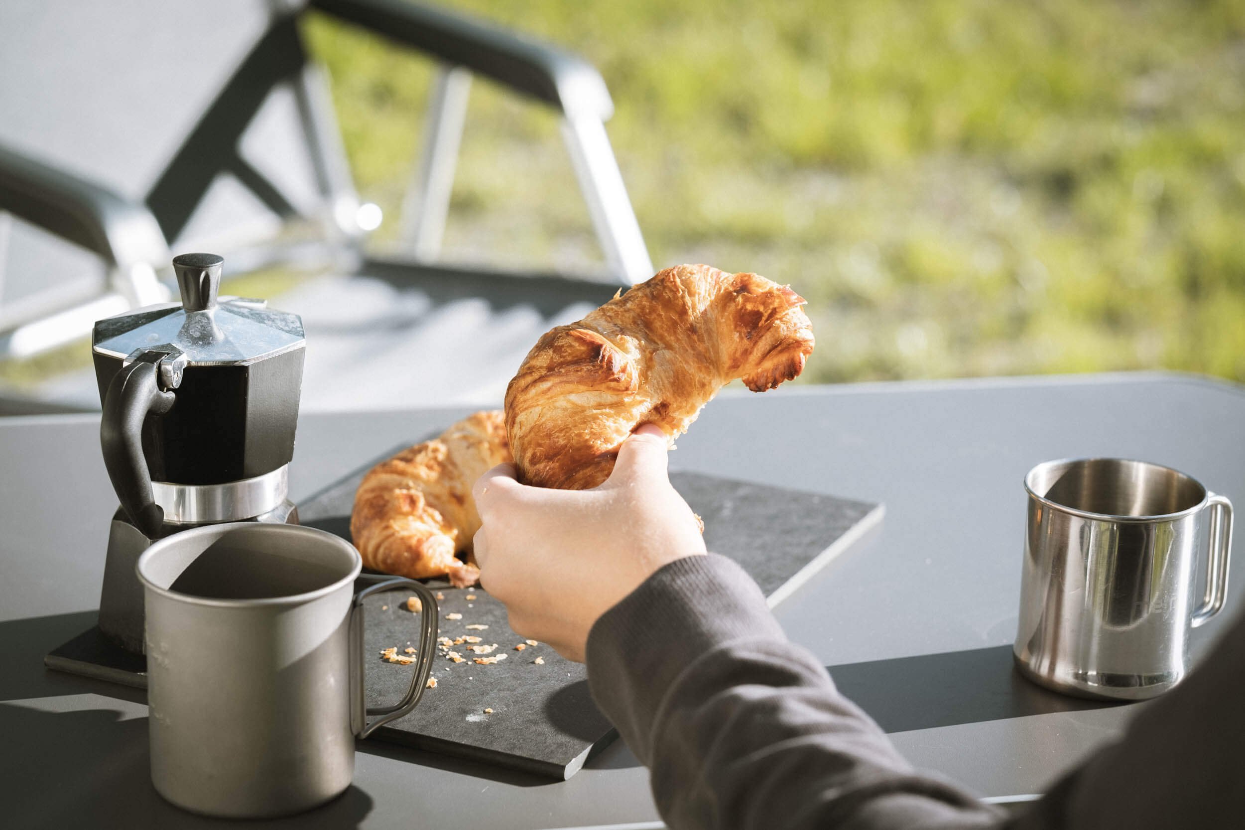 Eine Person hält ein Croissant über eine Küchentheke, auf der sich noch ein weiteres Croissant, eine Kaffeemaschine, eine graue Tasse und ein silberner Becher befinden. Draußen ist grüner Garten im Hintergrund sichtbar.