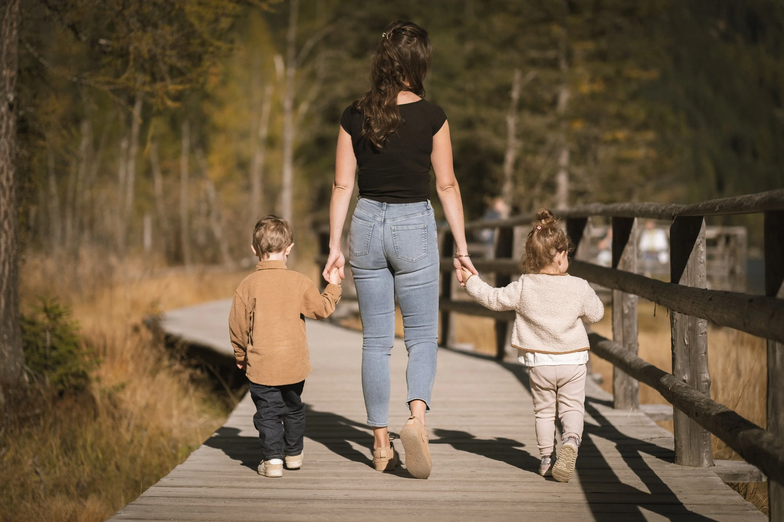 Eine Frau spaziert mit zwei kleinen Kindern auf einer Holzbrücke in einem Wald im Herbst. Alle sind von hinten zu sehen.