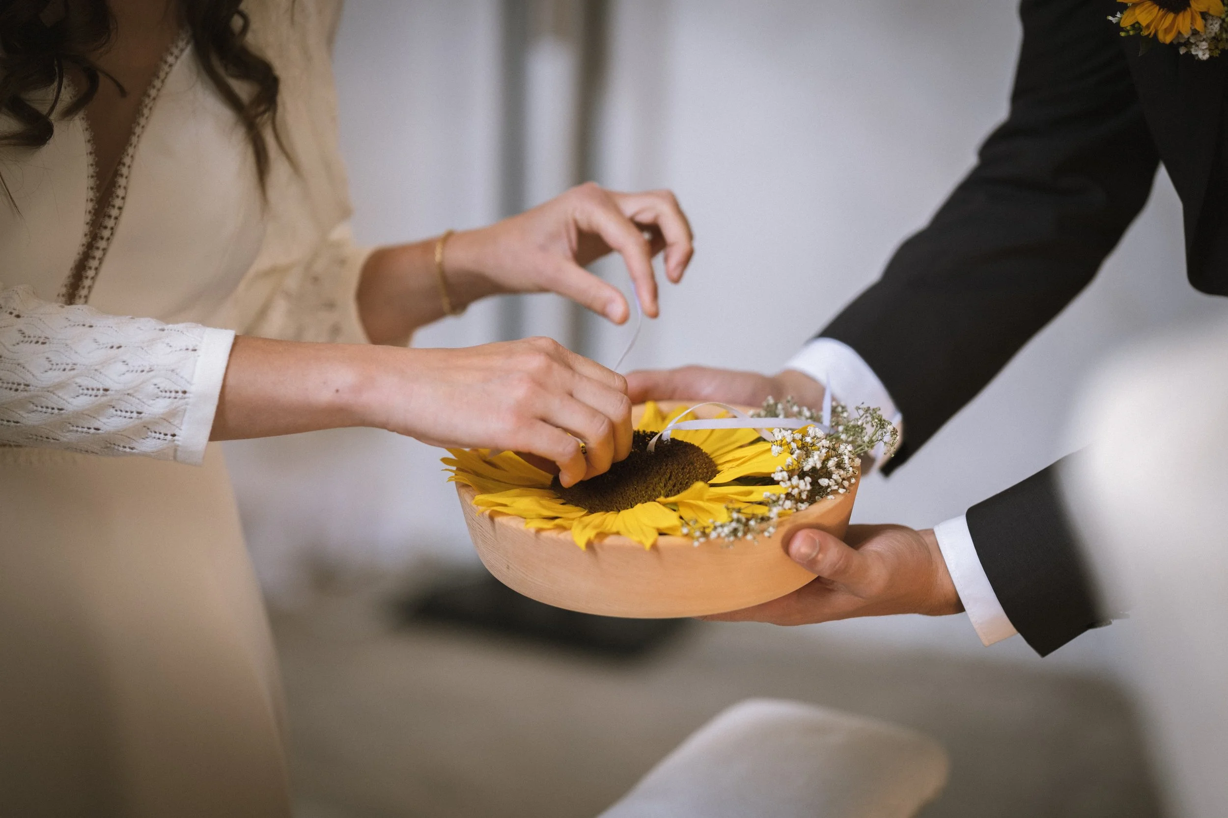 Zwei Personen bei einer Zeremonie, bei der eine Person einer anderen eine Sonnenblume in eine Schale legt, die mit Sonnenblumen und kleinen weißen Blumen dekoriert ist.