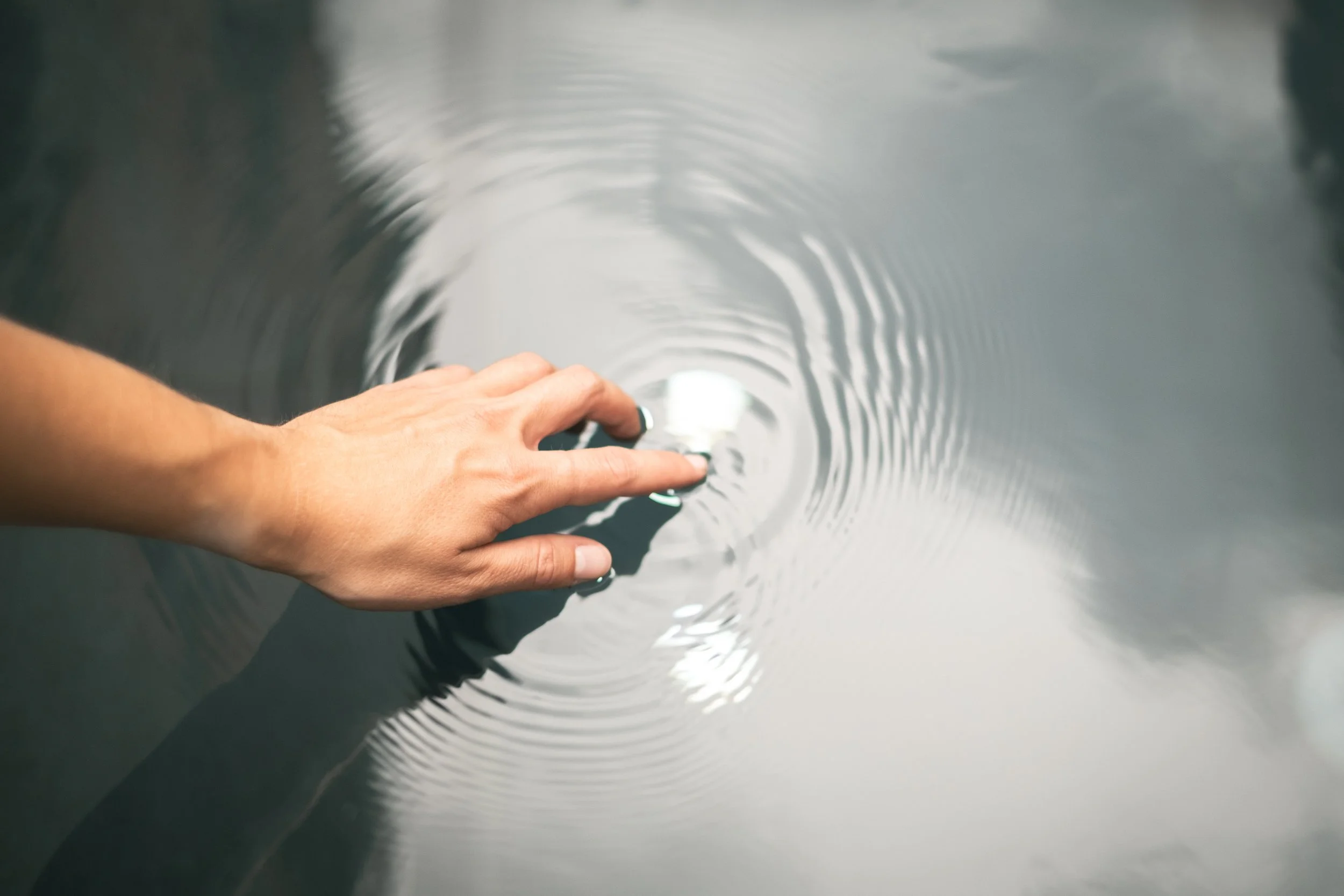 Eine Hand, die auf Wasser taucht und kleine Wellen erzeugt, während die Wasseroberfläche ruhig ist.