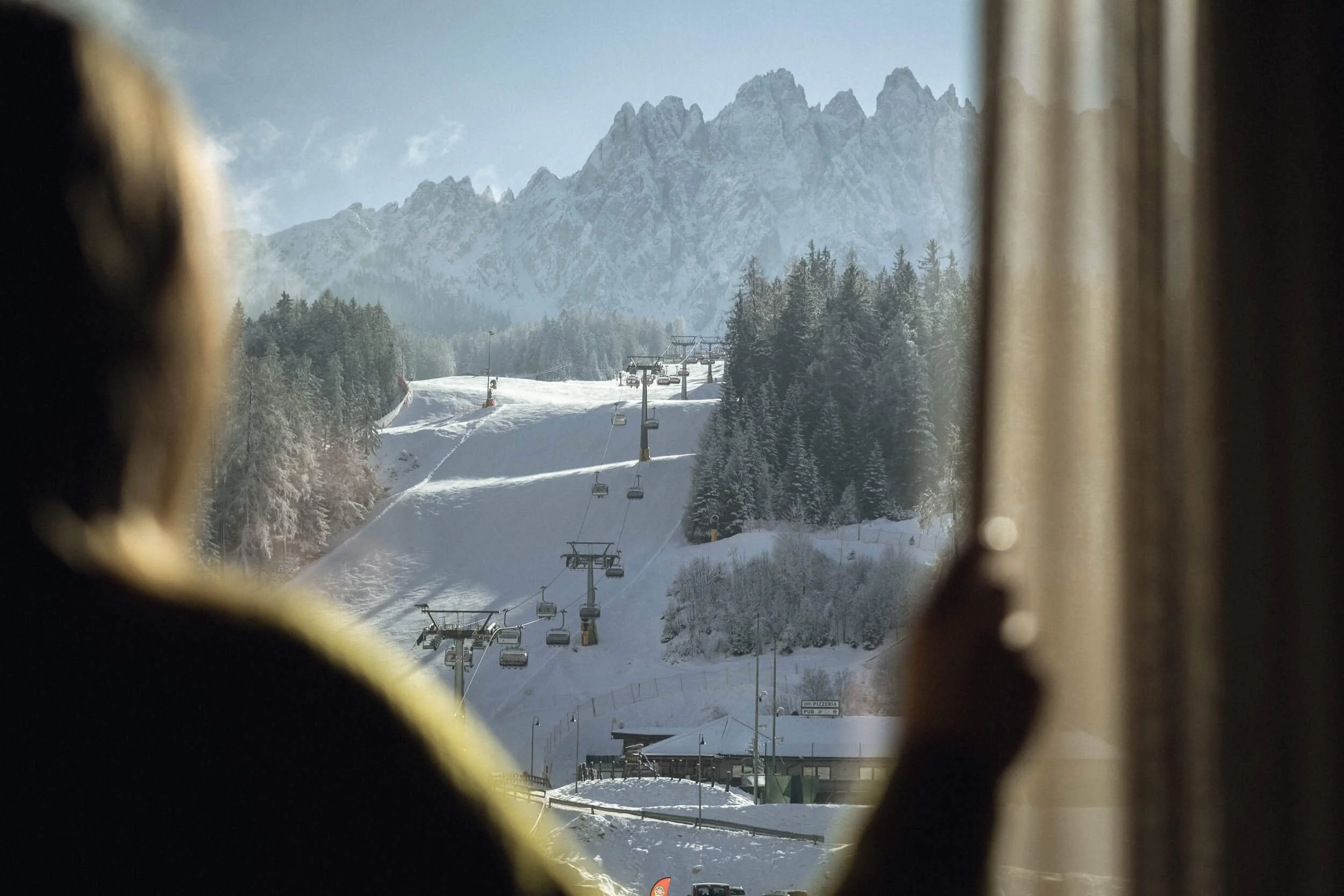 Eine Person schaut aus einem Fenster auf eine verschneite winterliche Berglandschaft mit Skiliften und schneebedeckten Bäumen