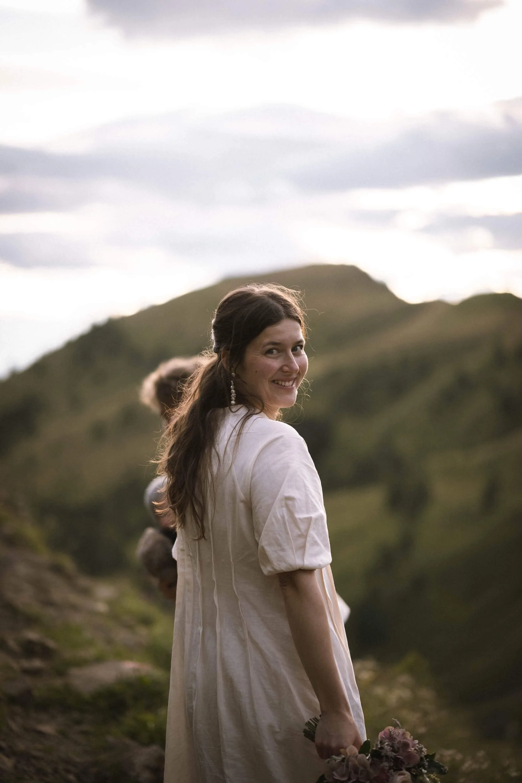 Eine lachende Frau in einem weißen Kleid, die auf einem Hügel in der Natur steht, im Hintergrund grüne Berge und ein bewölkter Himmel.