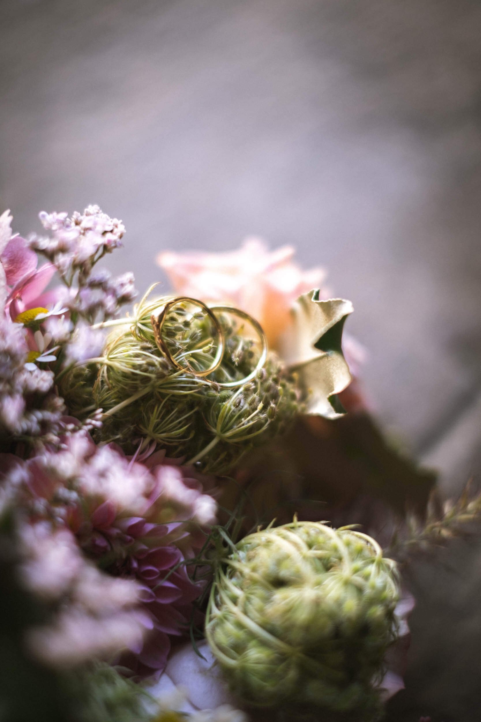 Zwei goldene Eheringe liegen auf einer Blumenarrangement aus pinken und weißen Blumen und grünen Pflanzen, mit unscharfem Hintergrund.