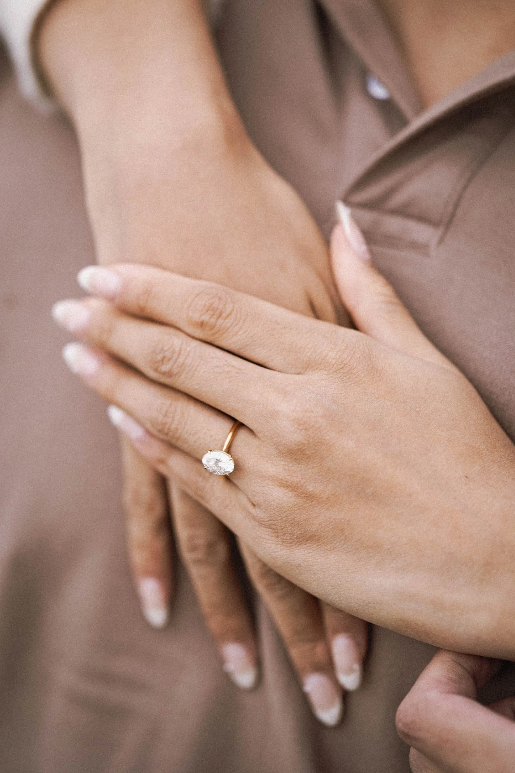 Nahaufnahme einer Hand mit einem Verlobungsring mit großem Diamanten, die auf die andere Hand gelegt ist, bei einer Person mit Nagellack in französischer Maniküre, im Hintergrund ein braunes Kleidungstück.