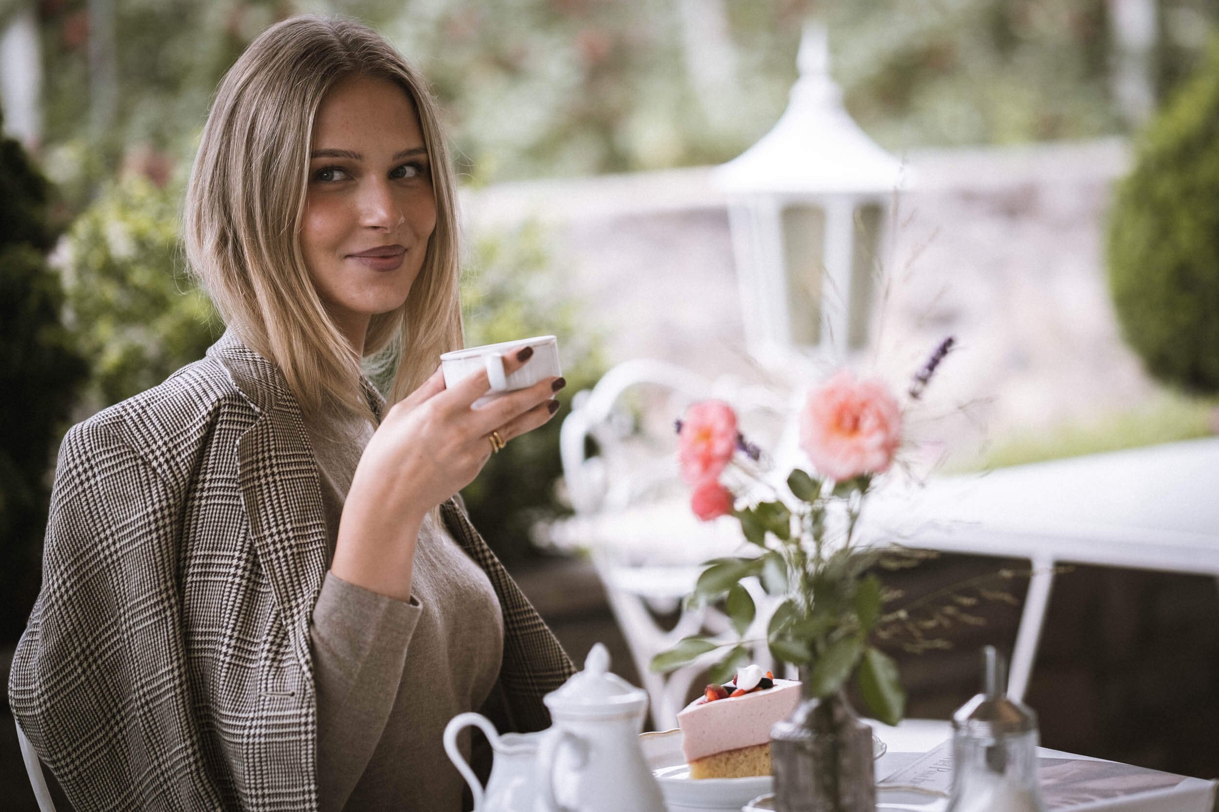 Junge Frau mit braunen Haaren, trägt karierte Jacke, sitzt bei einem Gartencafé und trinkt Kaffee, im Hintergrund Blumen und eine weiße Gartenlaube.