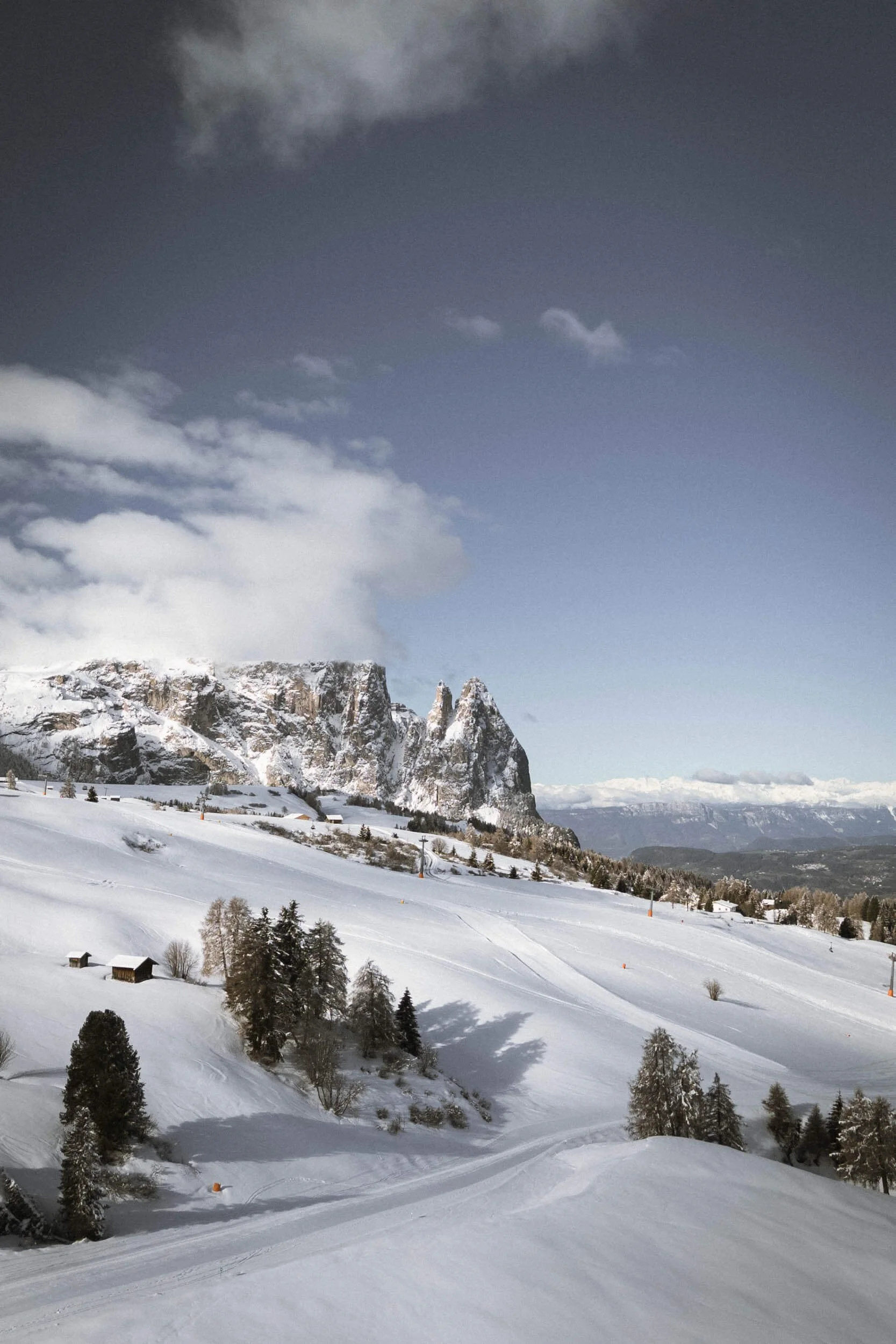 Schneebedeckte Berge und Hänge unter einem bewölkten Himmel, mit kleinen Hütten und Bäumen im Vordergrund.