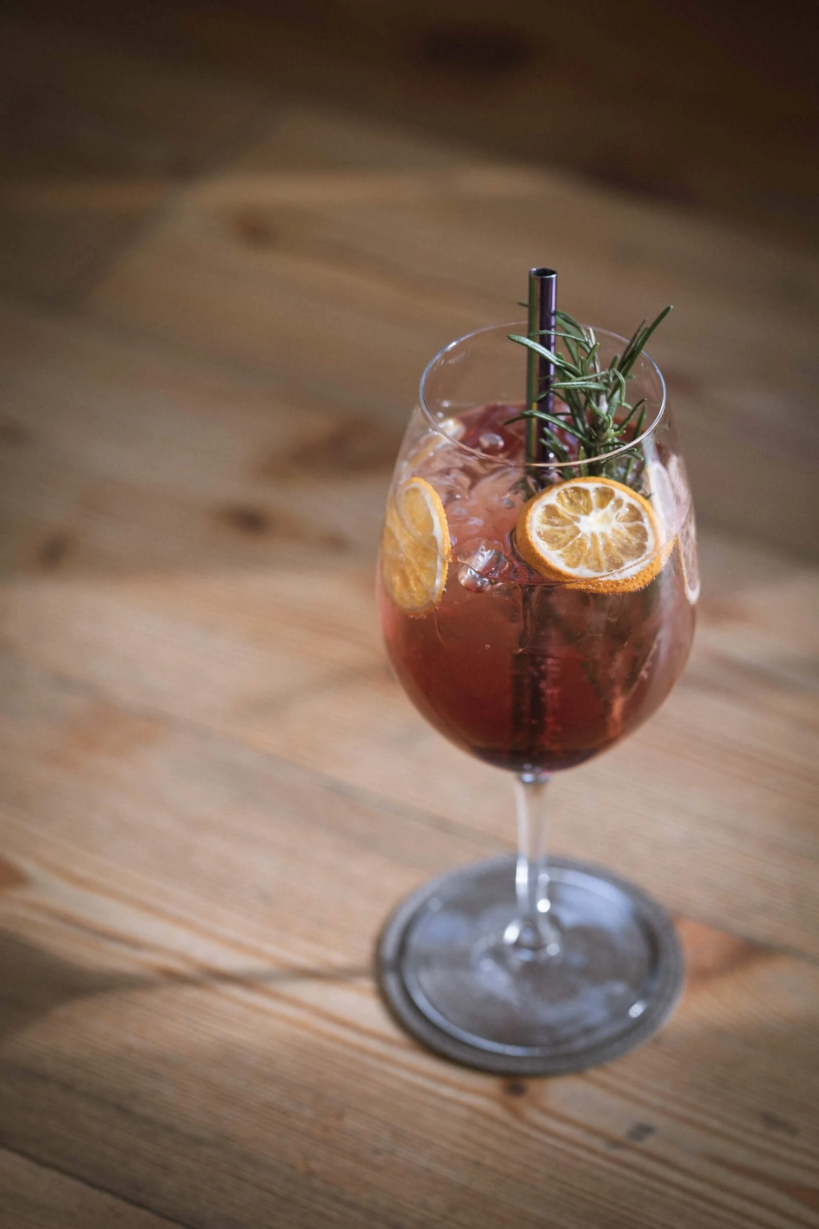 Ein bunter Sommerdrink in einem Weinglas mit Zitronenscheiben, Rosmarinzweig, Strohhalm und Eiswürfeln auf einem Holztisch.